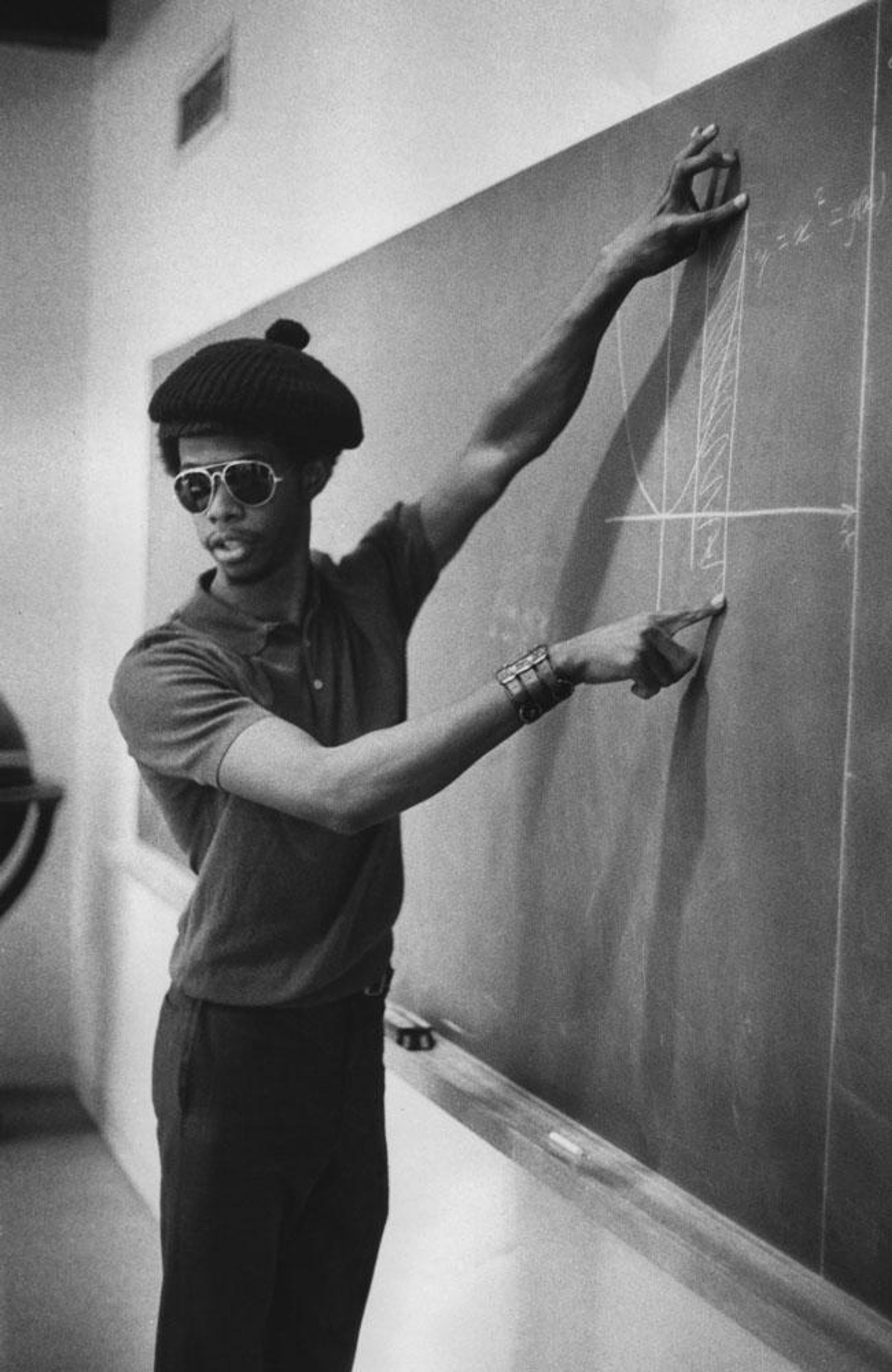 A young man in sunglasses and a black beret points towards a scientific diagram on a blackboard.
