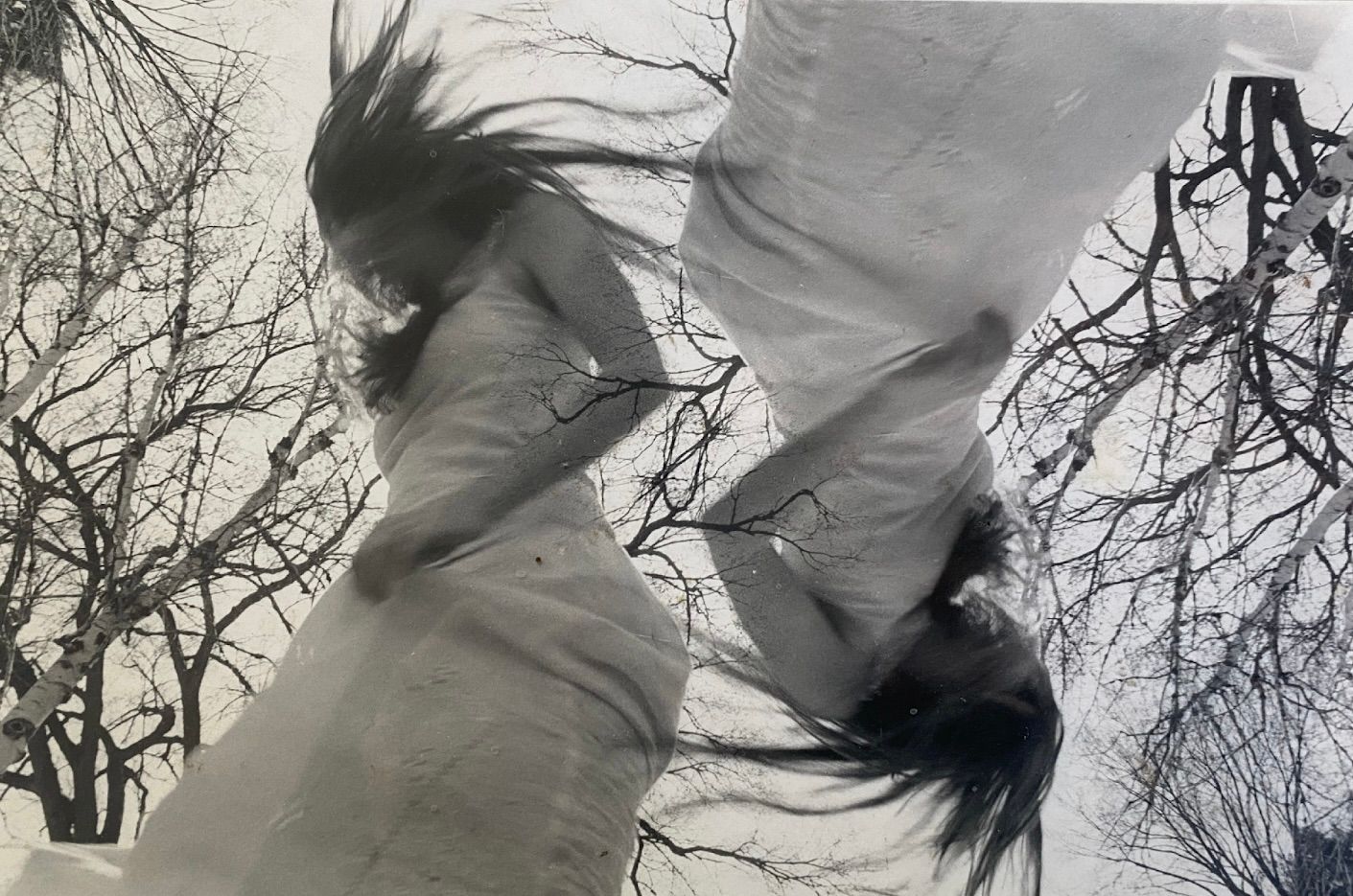 A black and white image of two inverted twirling figures against the backdrop of trees and sky. 