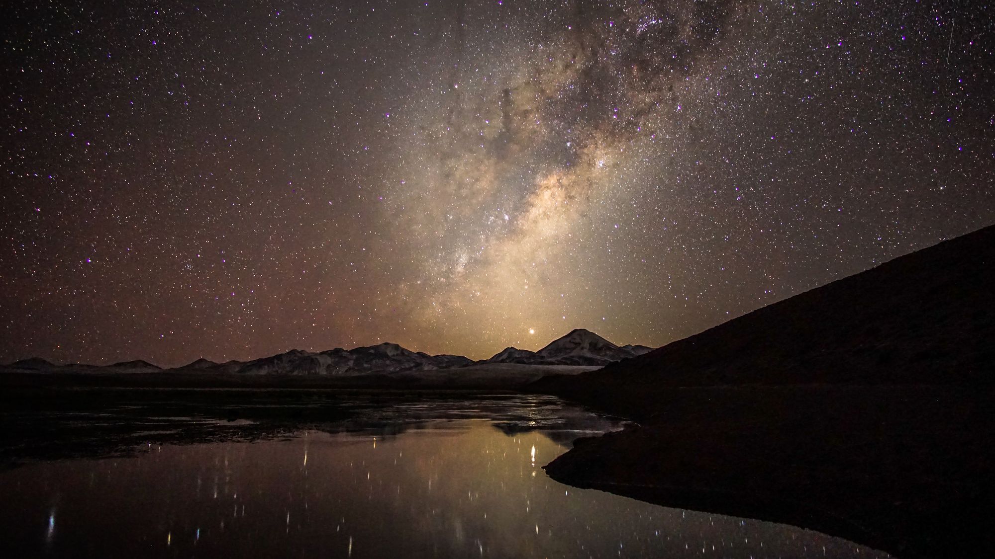 A starry night with the milky way glowing above a body of water, with a mountain range in the background.