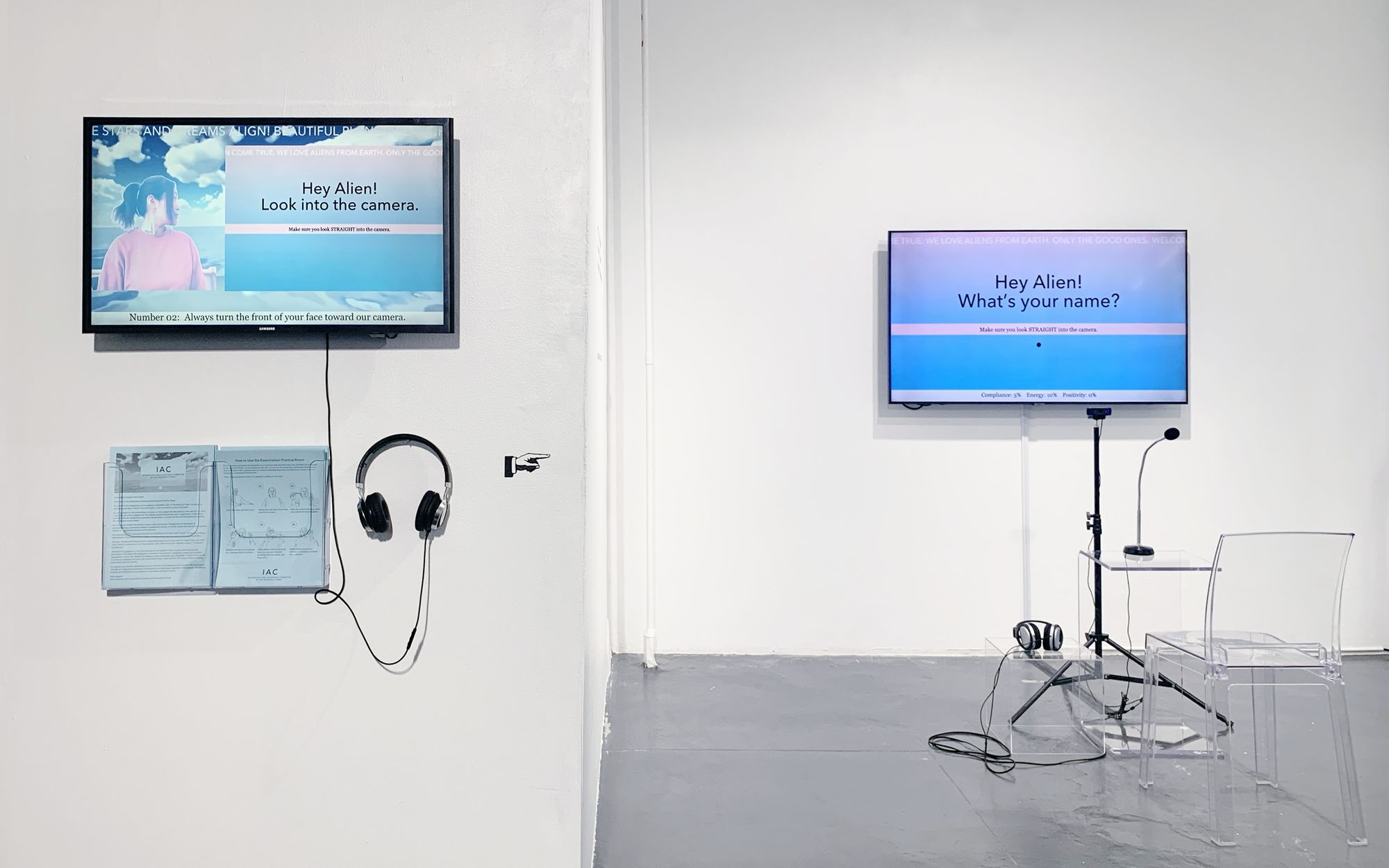 A monitor positioned on a blank white wall reads "Hey Alien" in pastel colors, with a headset hung directly below it.