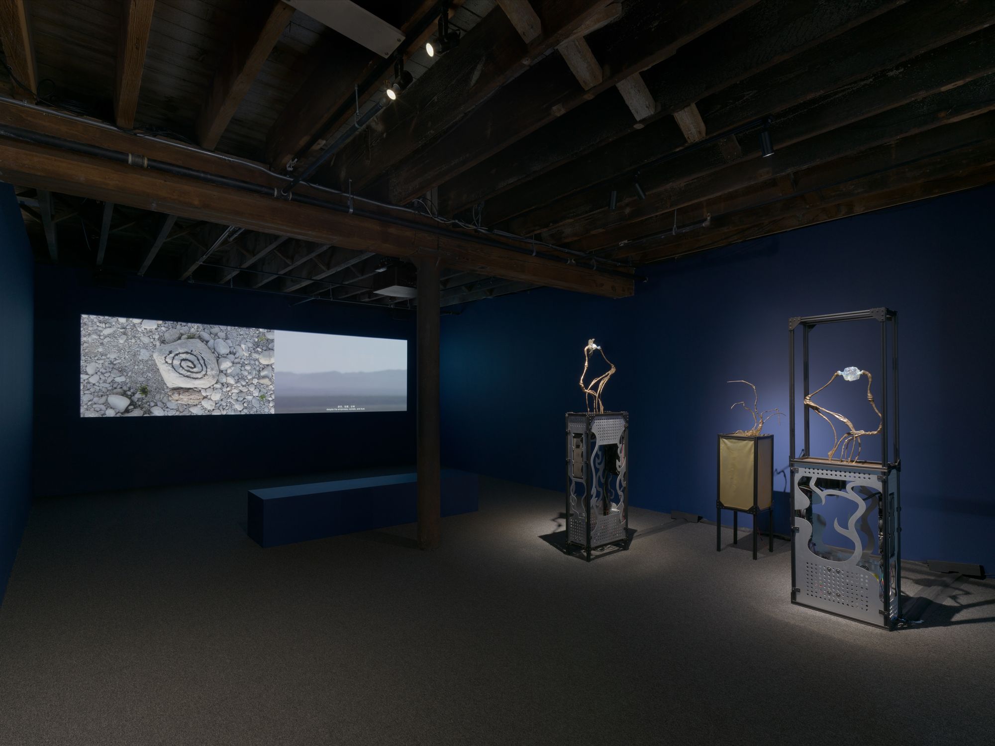 A dark gallery, with a projected spiral image in the corner and other metal-like objects close by.