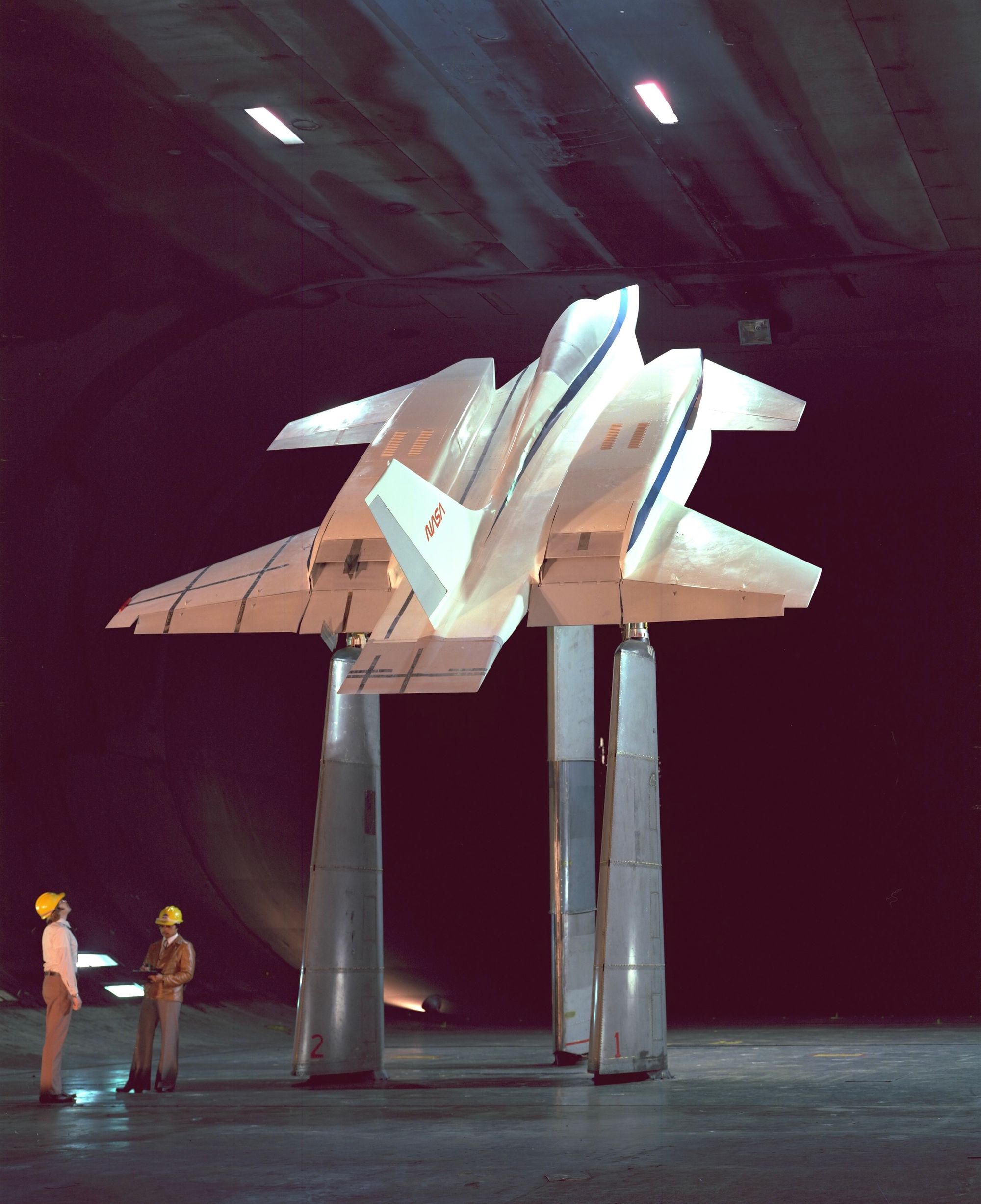 Three figures in lab coats and yellow construction hats look upwards at a large yellow helicopter-like object.