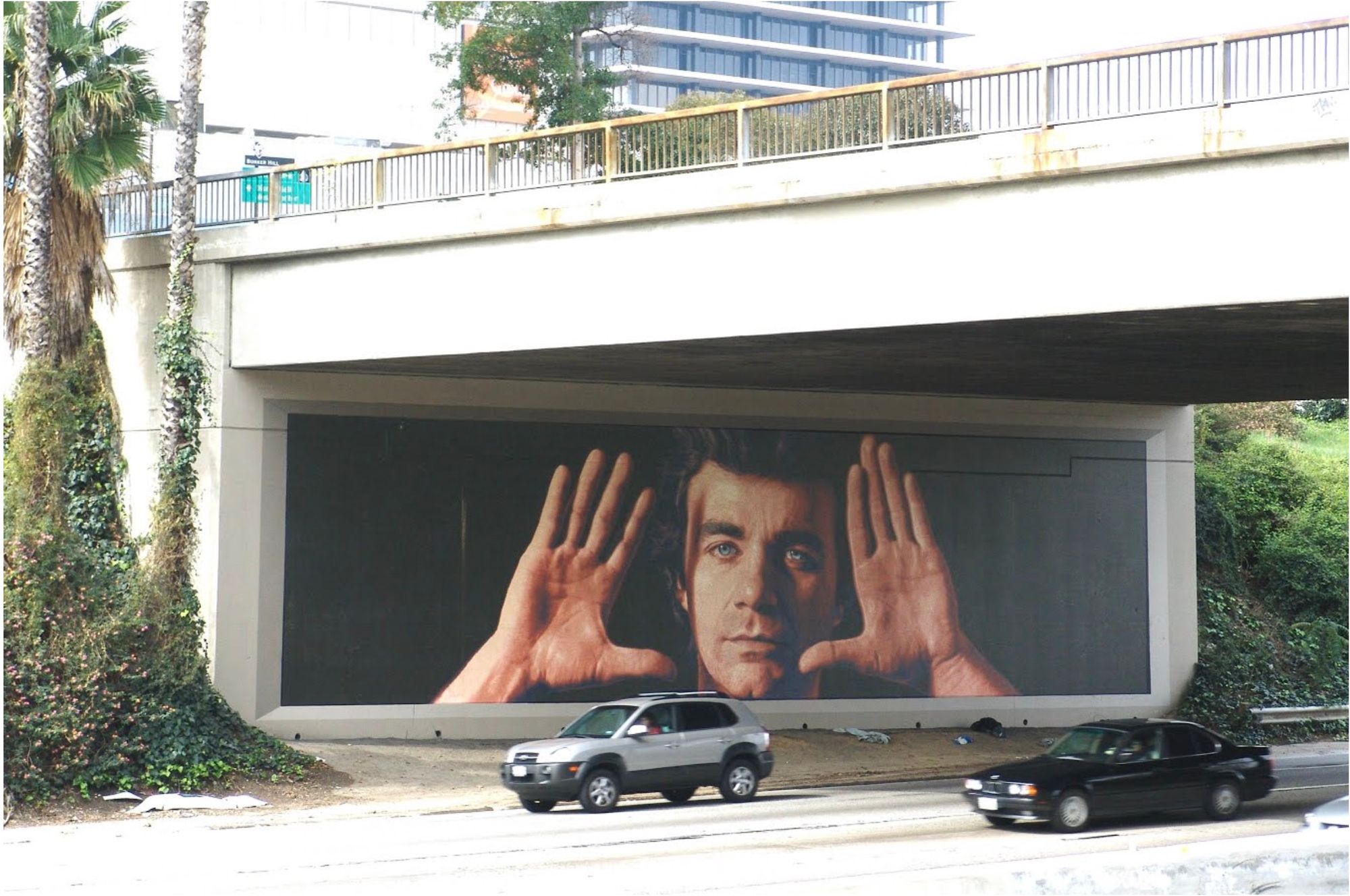 A mural on the side of the freeway featuring a man peering out from between his hands.