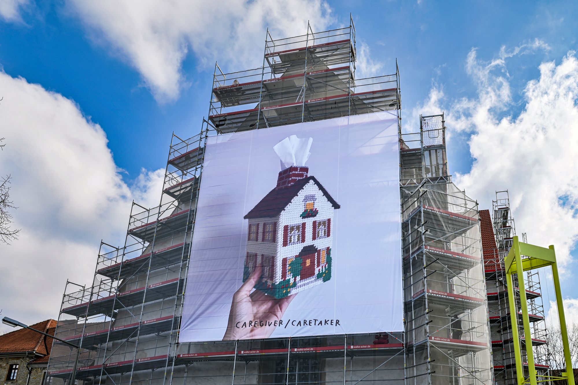 Billboard with white person's hand holding a crocheted house tissue box with the words "Caretaker/Caregiver" on the side of scaffolding. 