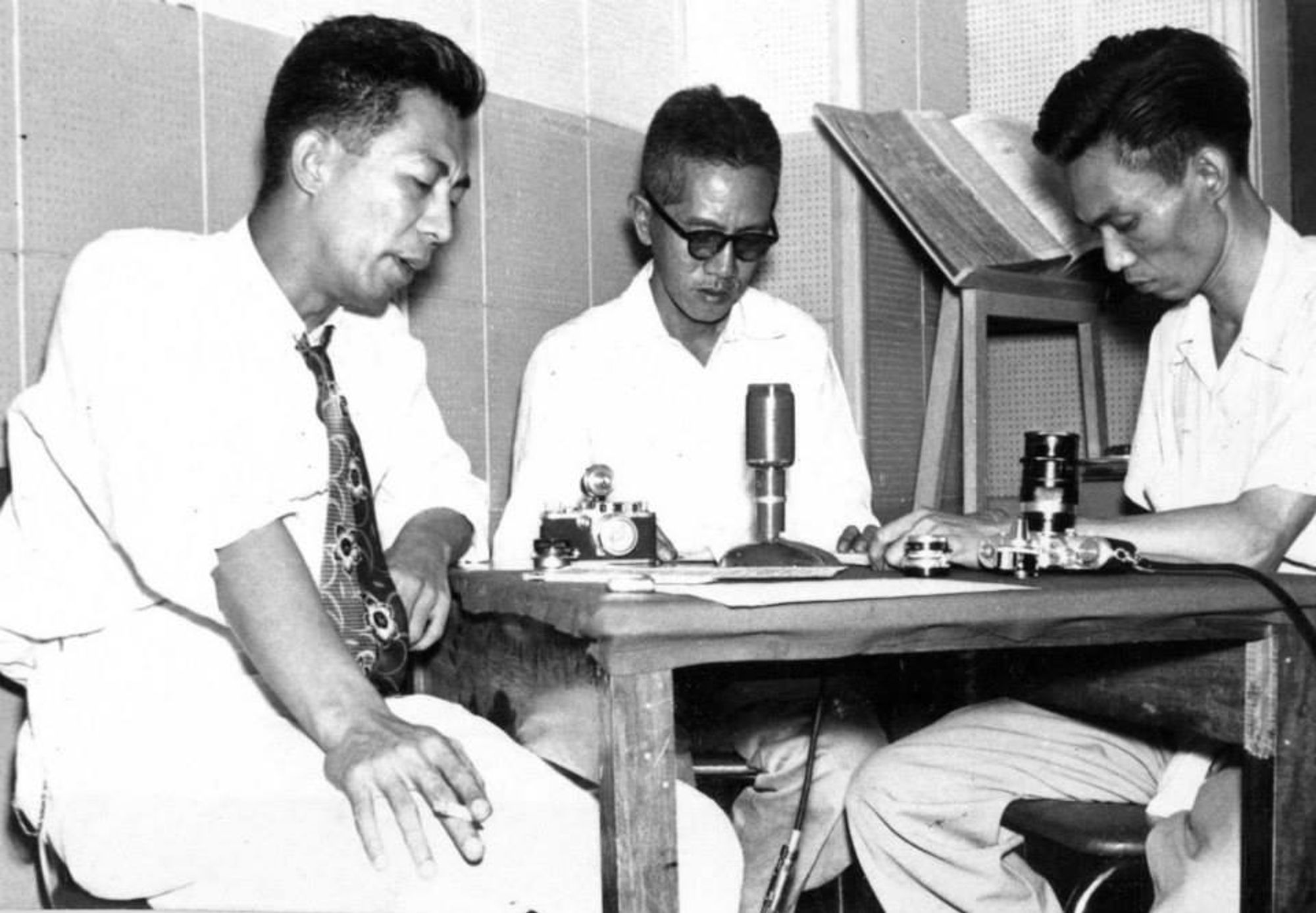 A black-and-white photograph of three photographers, including Protomartir, sitting at a table with cameras on it. They are all looking down at the table with concentration and one of them is holding a cigarette in his hand.