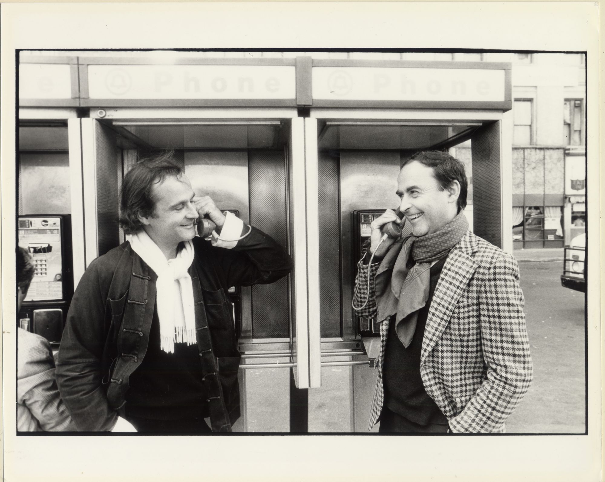 Two men in a phone booth face each other while talking.
