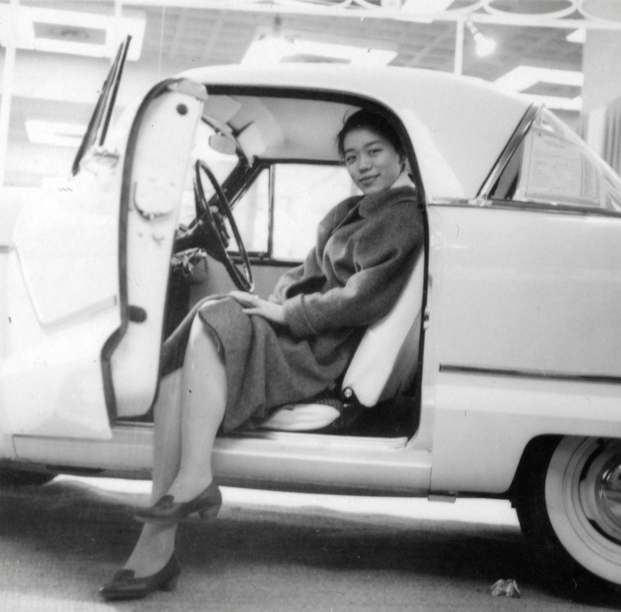 A woman sitting in a white car, her legs resting on the ground outside