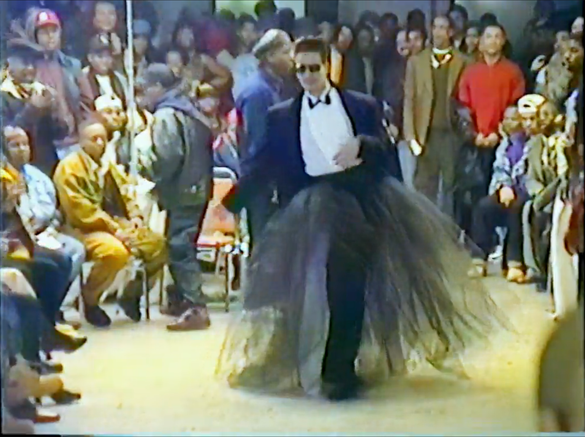 A man wearing a suit with a skirt and sunglasses walking across a ballroom stage 