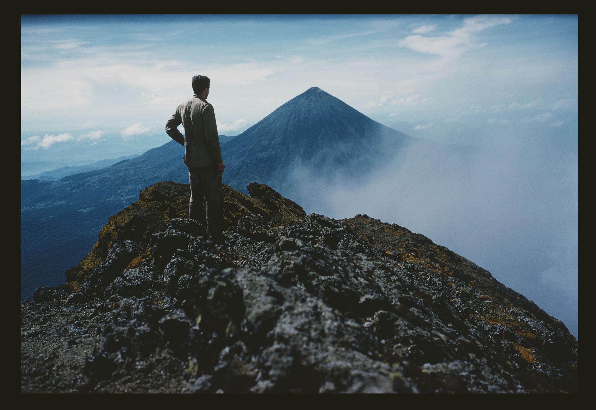 A man stands at the top of a mountain, surrounded by fog and neighboring peaks.
