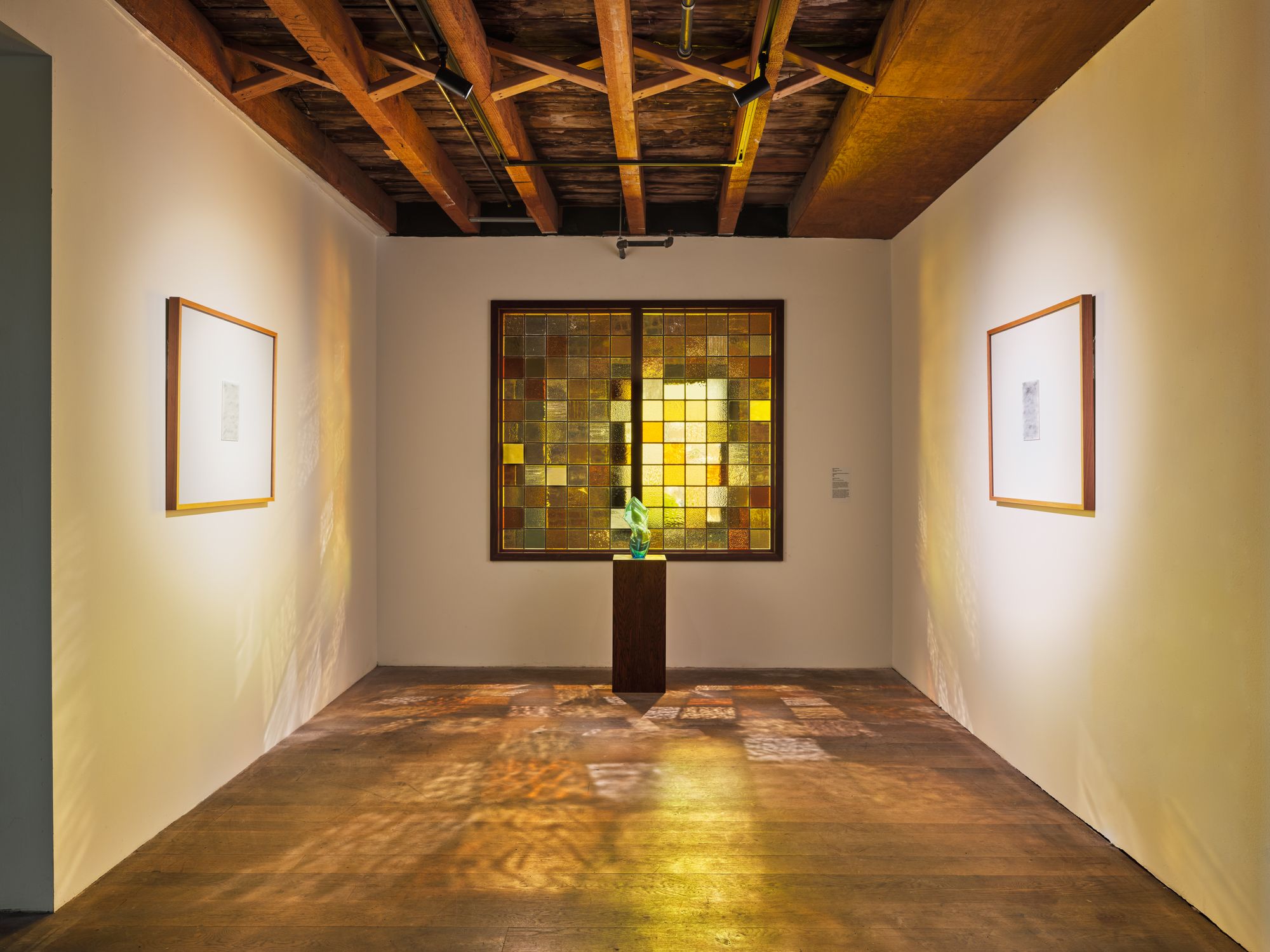 A gallery washed in yellow light from a stained glass window against the back wall, featuring a small podium holding a glass sculpture and two paintings on both side walls. 