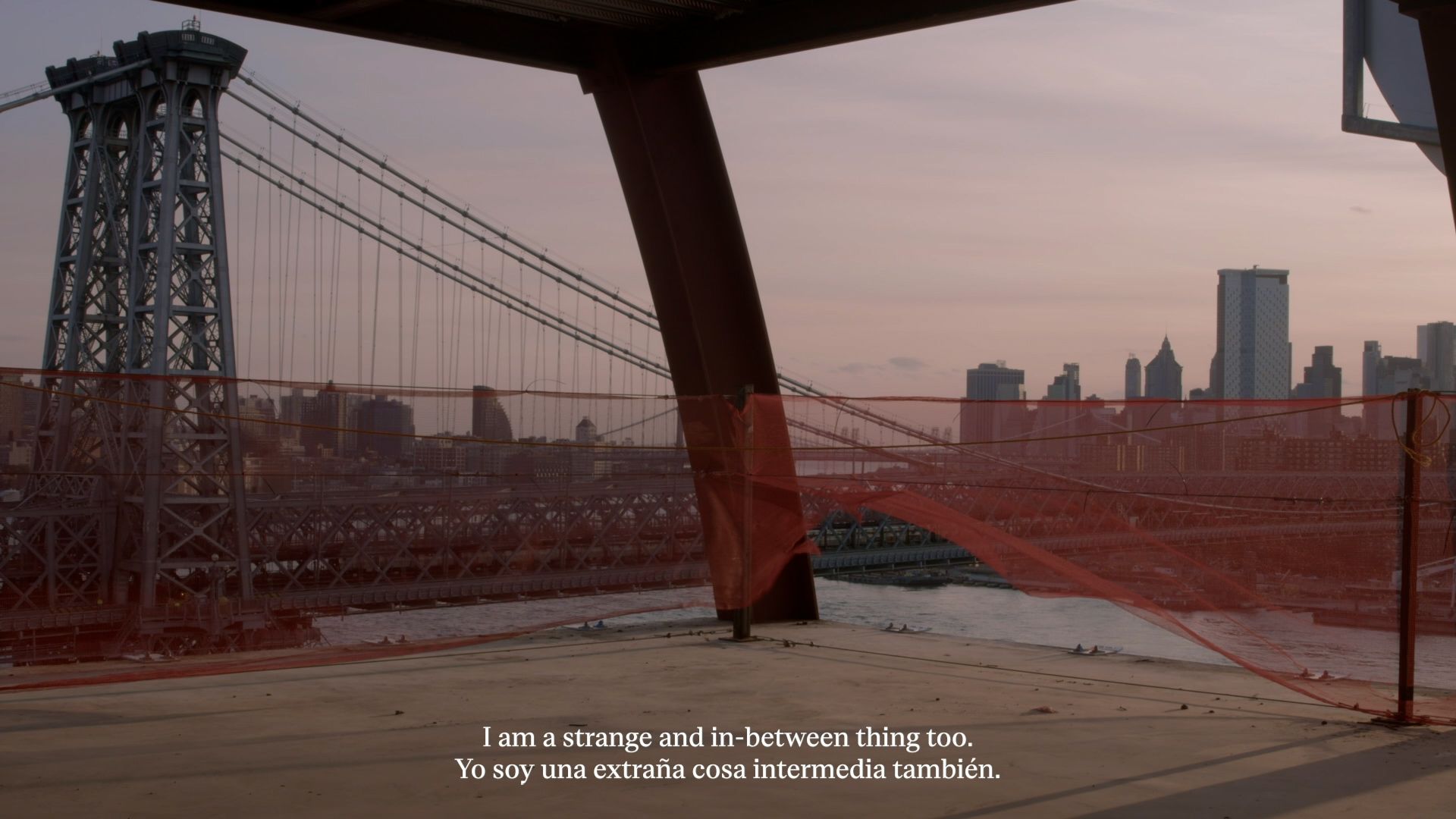 A film still that looks upon the Williamsburg Bridge from behind the orange gauze of a construction site, with a caption in white text at the bottom of the screen which reads: "I am a strange and in-between thing too" in both english and spanish.