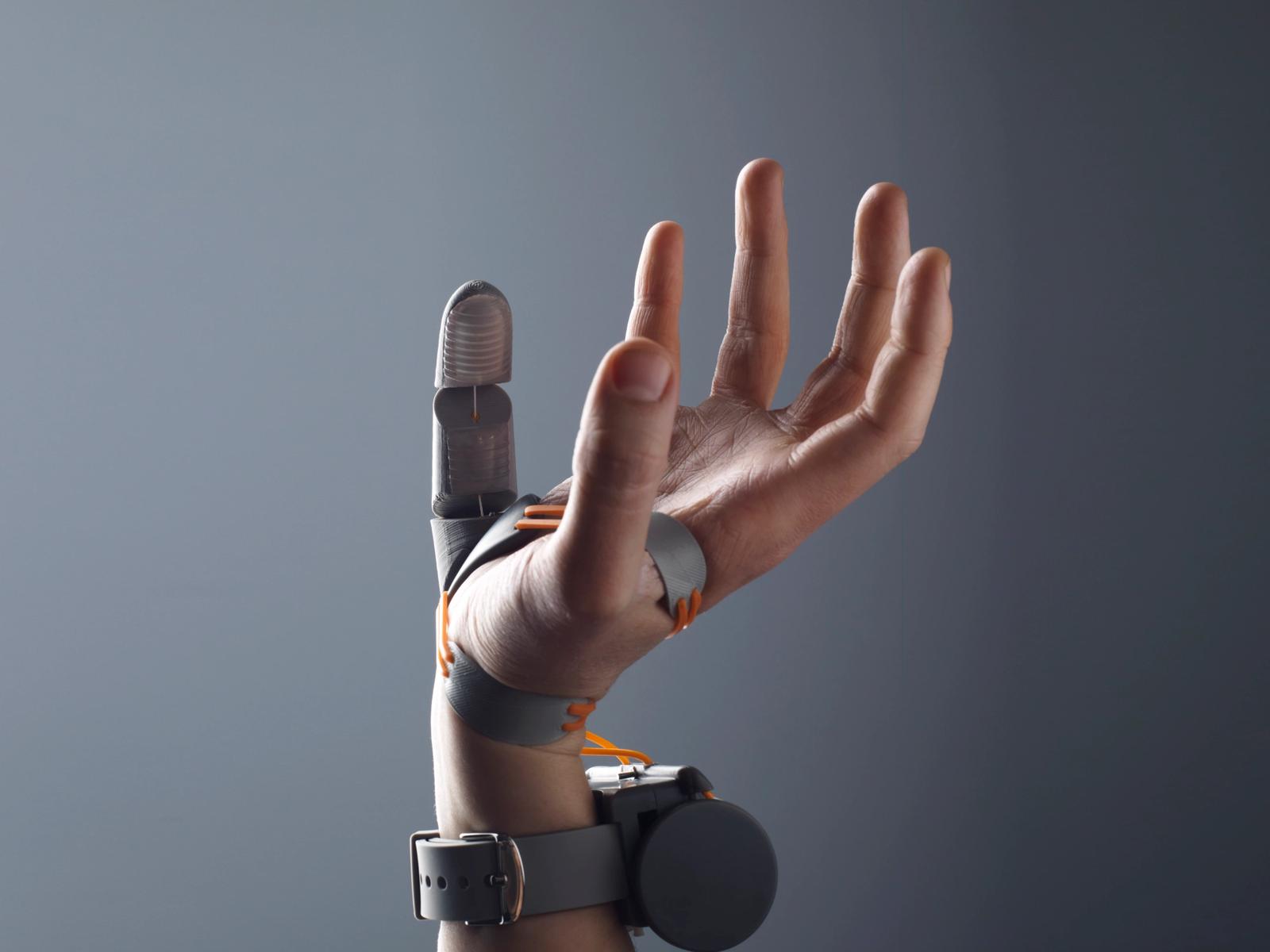 A prosthetic thumb attachment is affixed to a hand that is raised in the air. The background is grey and there is a little bit of light coming from the top left side of the image.