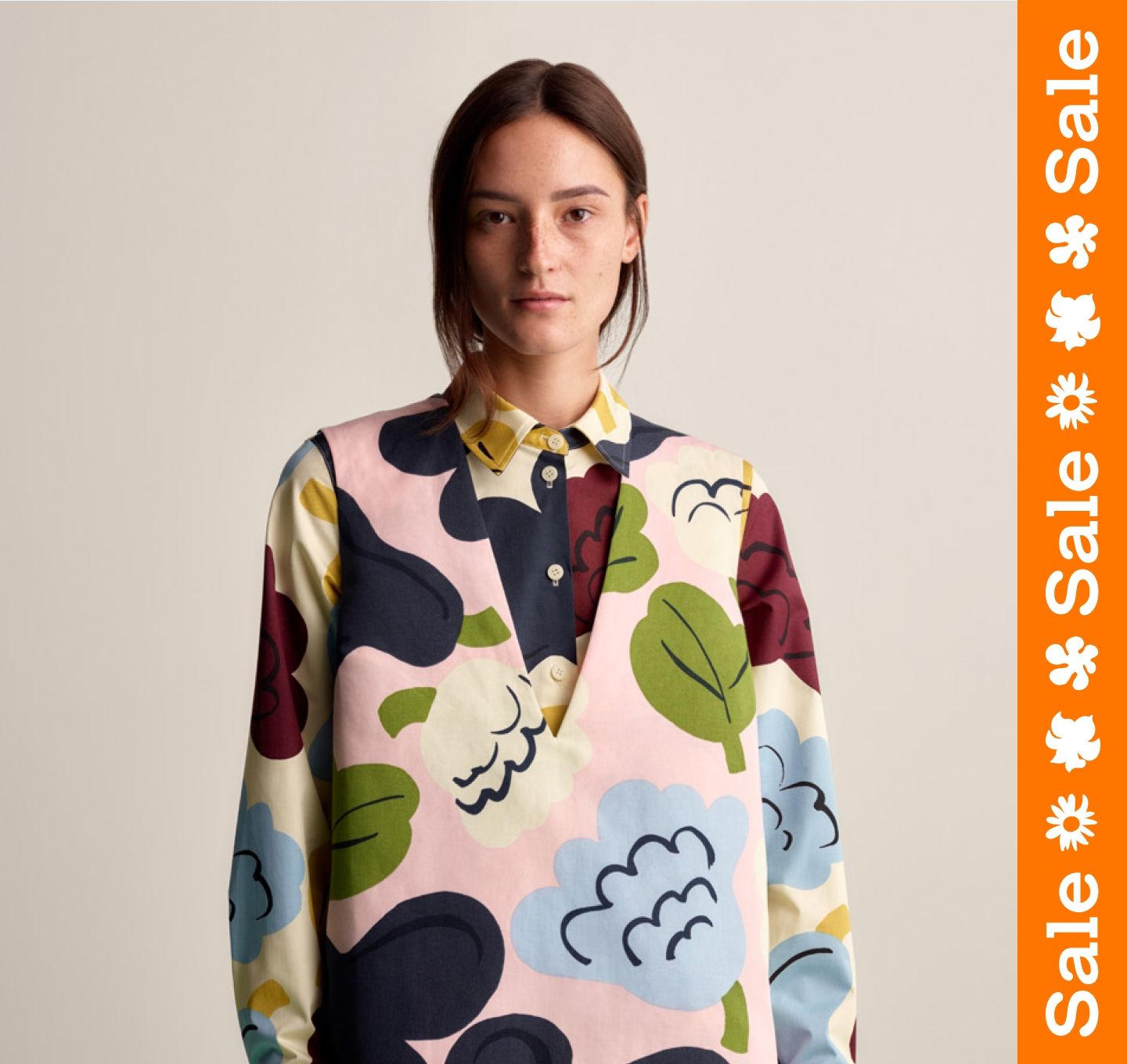 Model wearing a sleeveless dress in Kukasta Kukkaan print, and a collar shirt under in the same print. Orange sale stripe on the right.