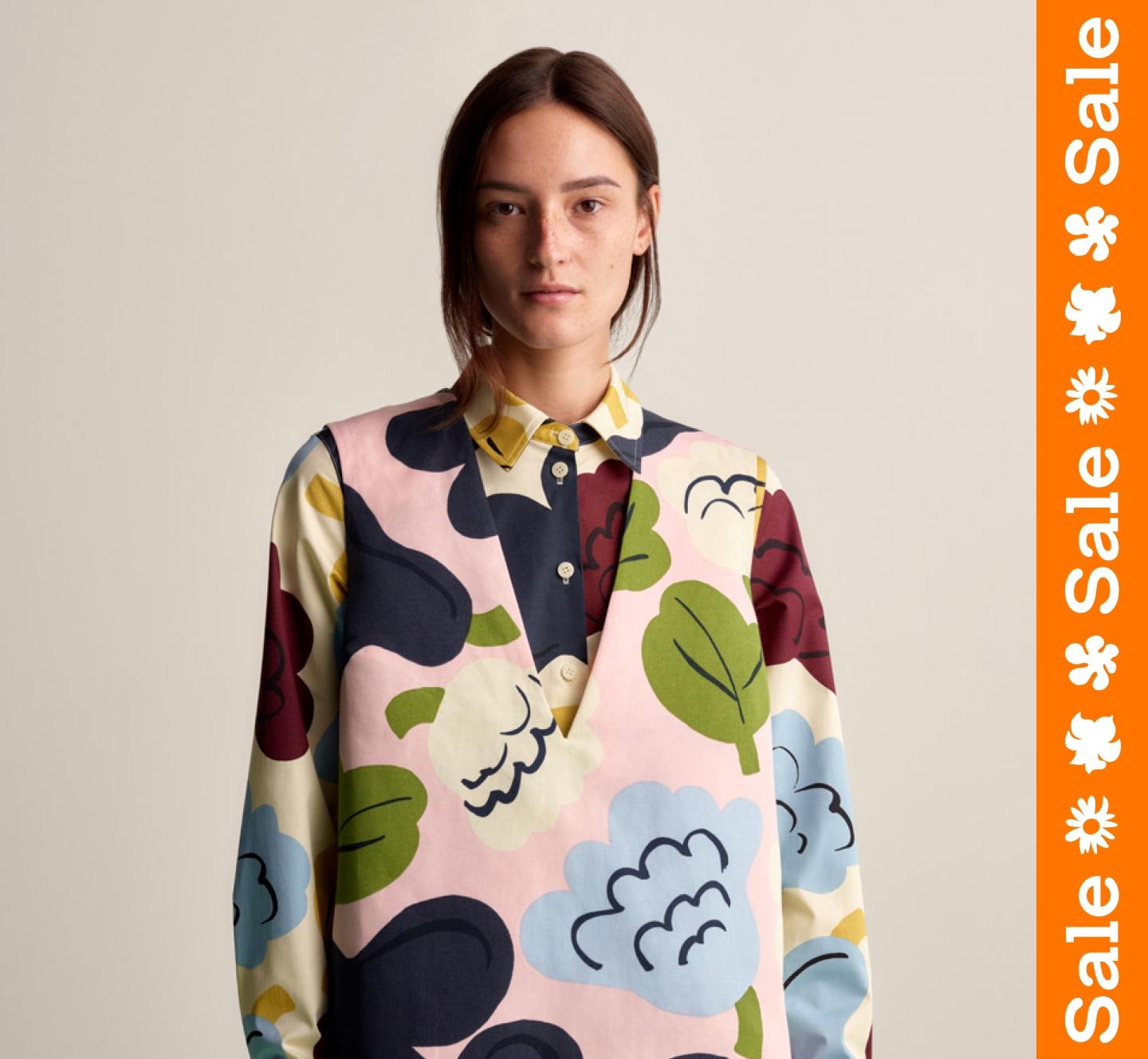 Model wearing a sleeveless dress in Kukasta Kukkaan print, and a collar shirt under in the same print. Orange sale stripe on the right.