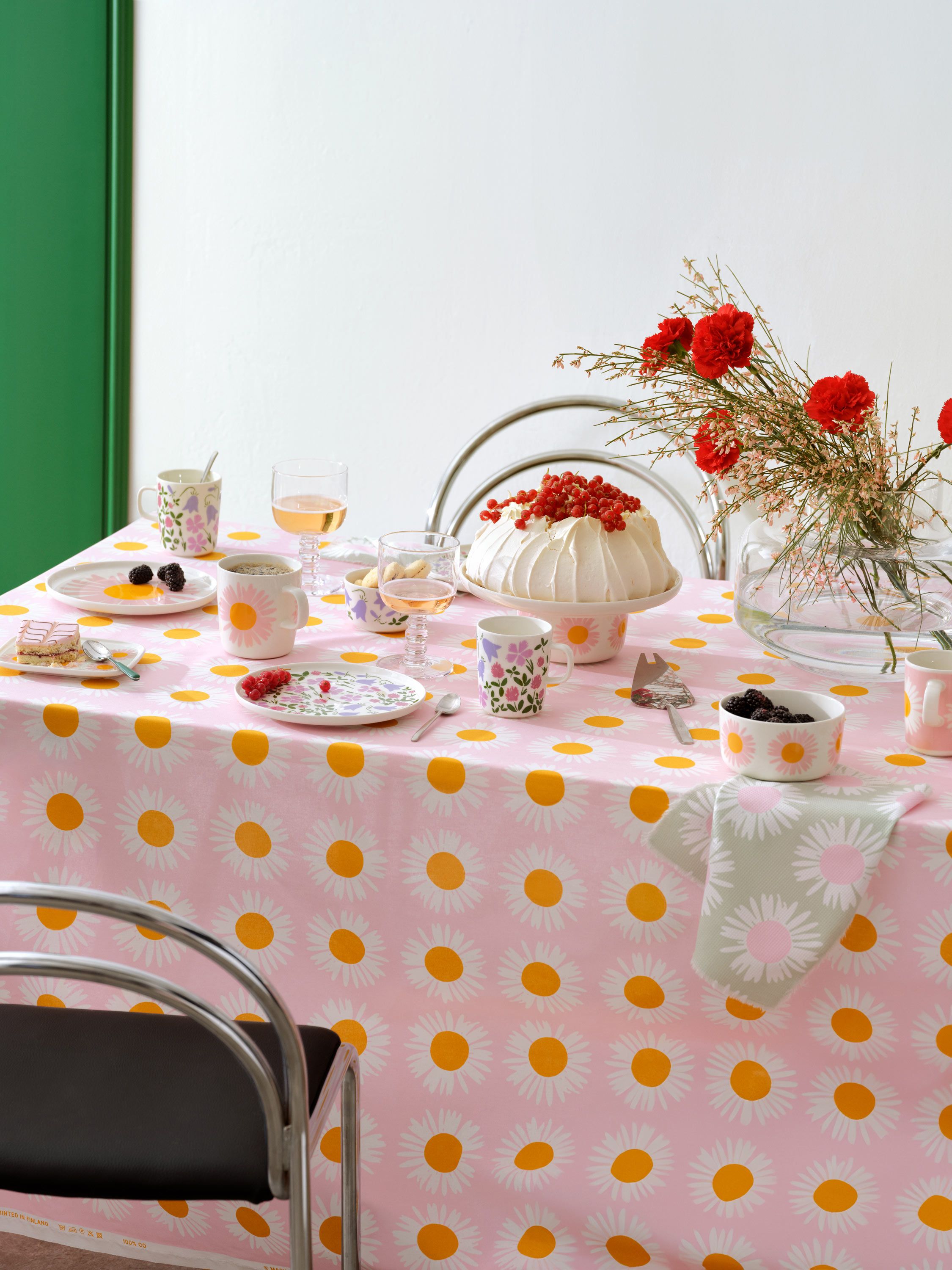 Katettu pöytä, jossa on vaaleanpunainen Auringonkukka-pöytäliina, kakku ja Sisilisko-astioita.