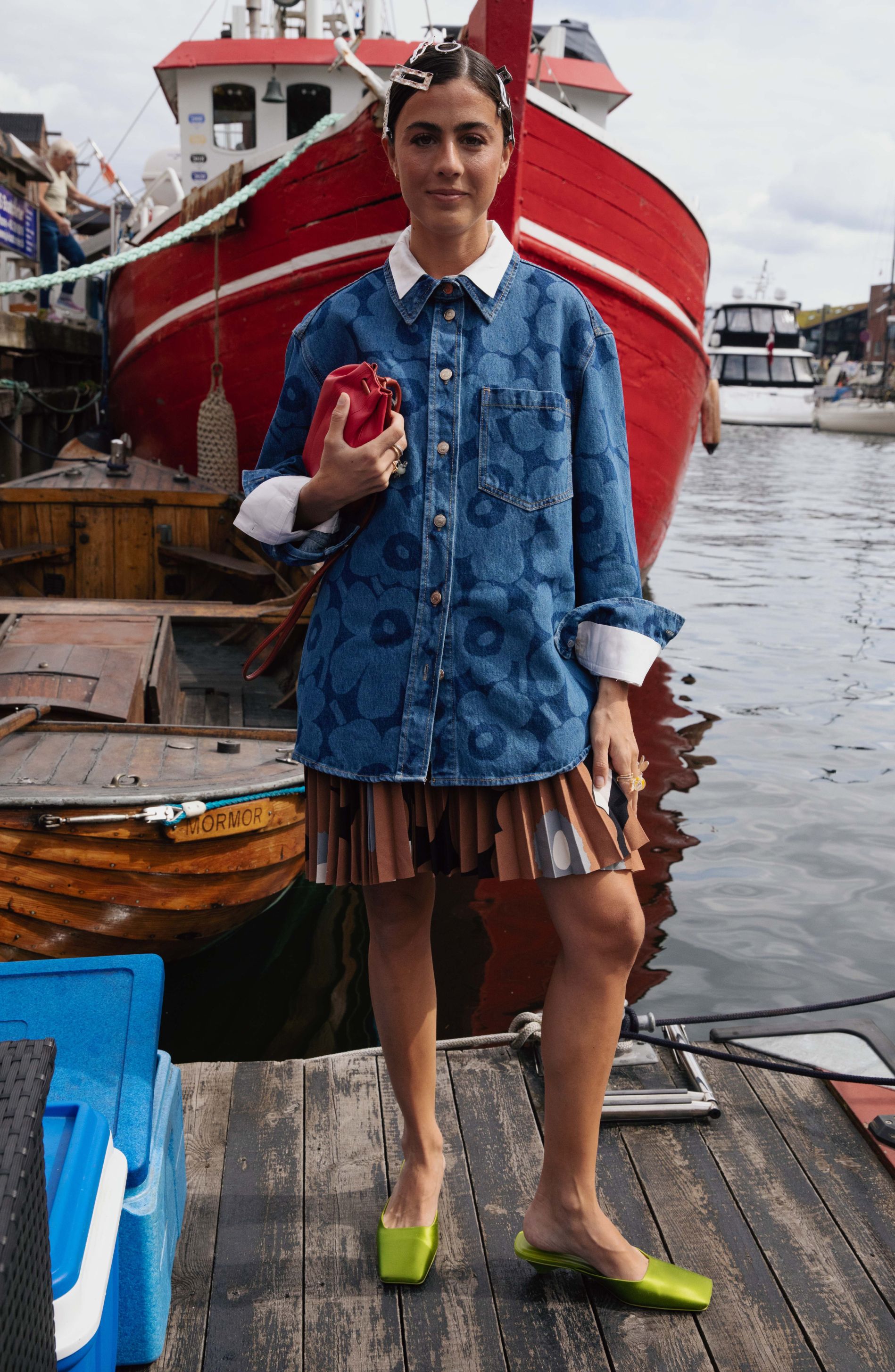 Vicky Montanari stands next to a boat wearing a Unikko denim jacket and a pleated skirt in the Unikko pattern