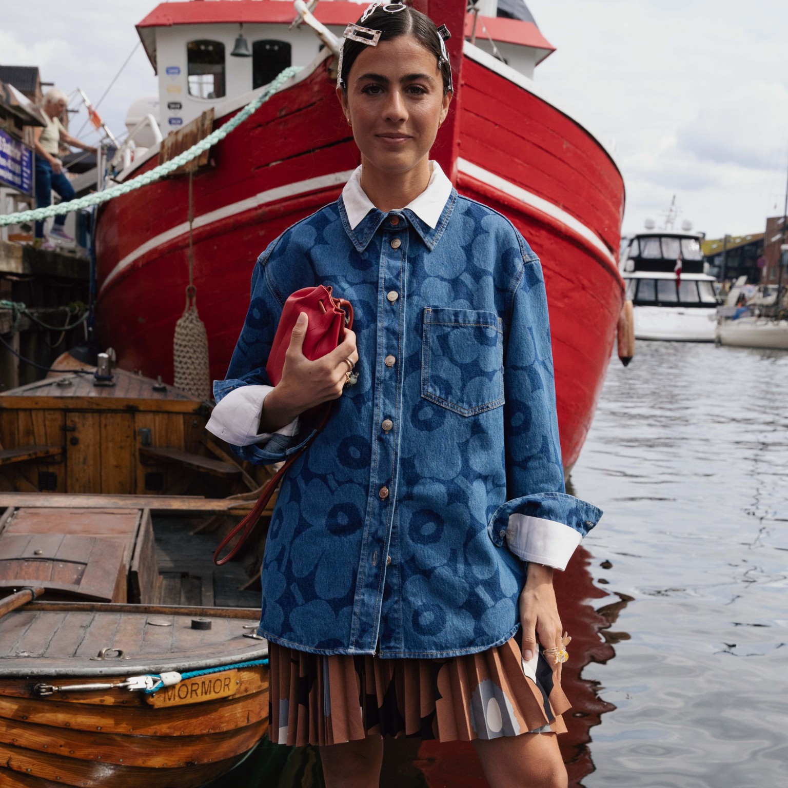 Vicky Montanari stands next to a boat wearing a Unikko denim jacket and a pleated skirt in the Unikko pattern