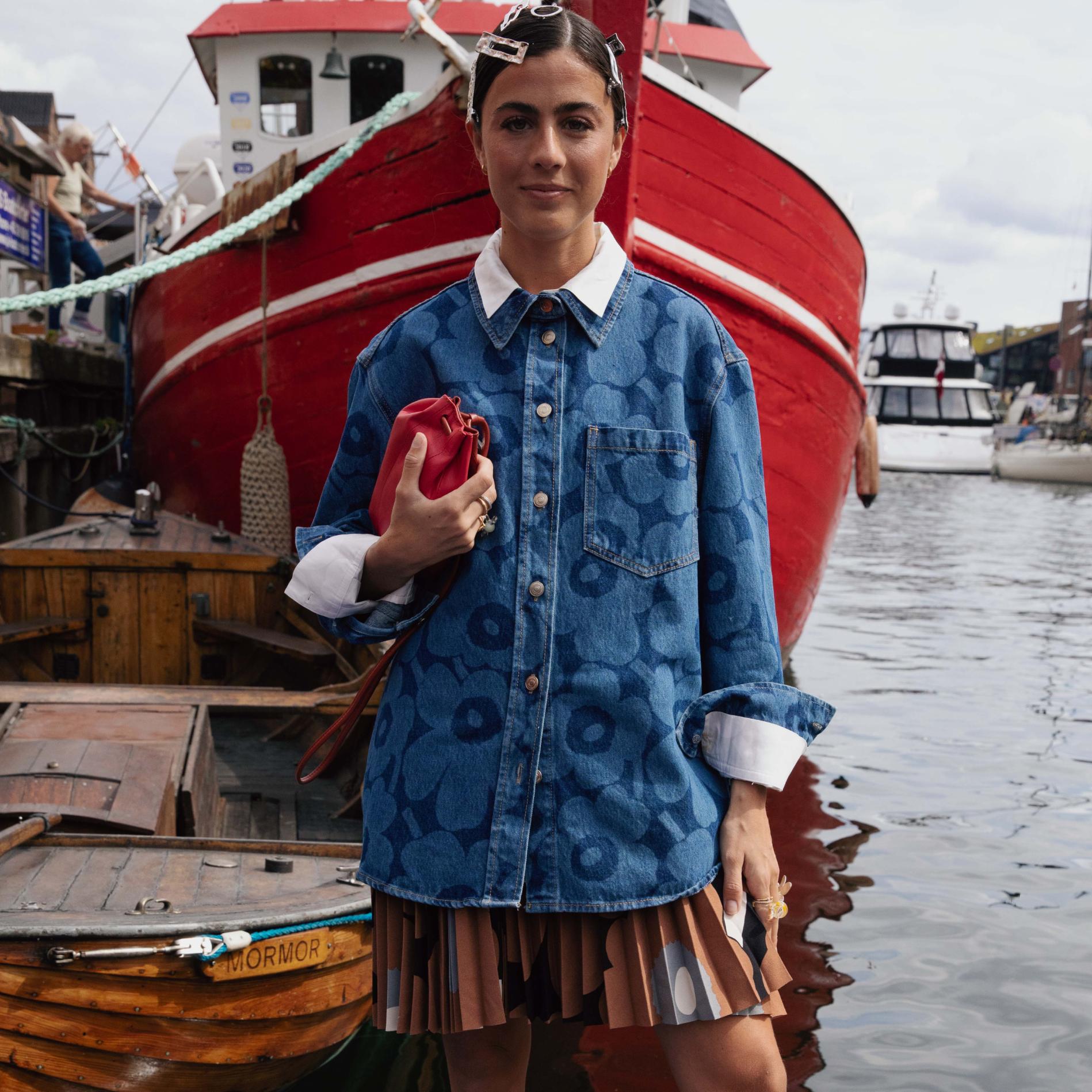 Vicky Montanari stands next to a boat wearing a Unikko denim jacket and a pleated skirt in the Unikko pattern