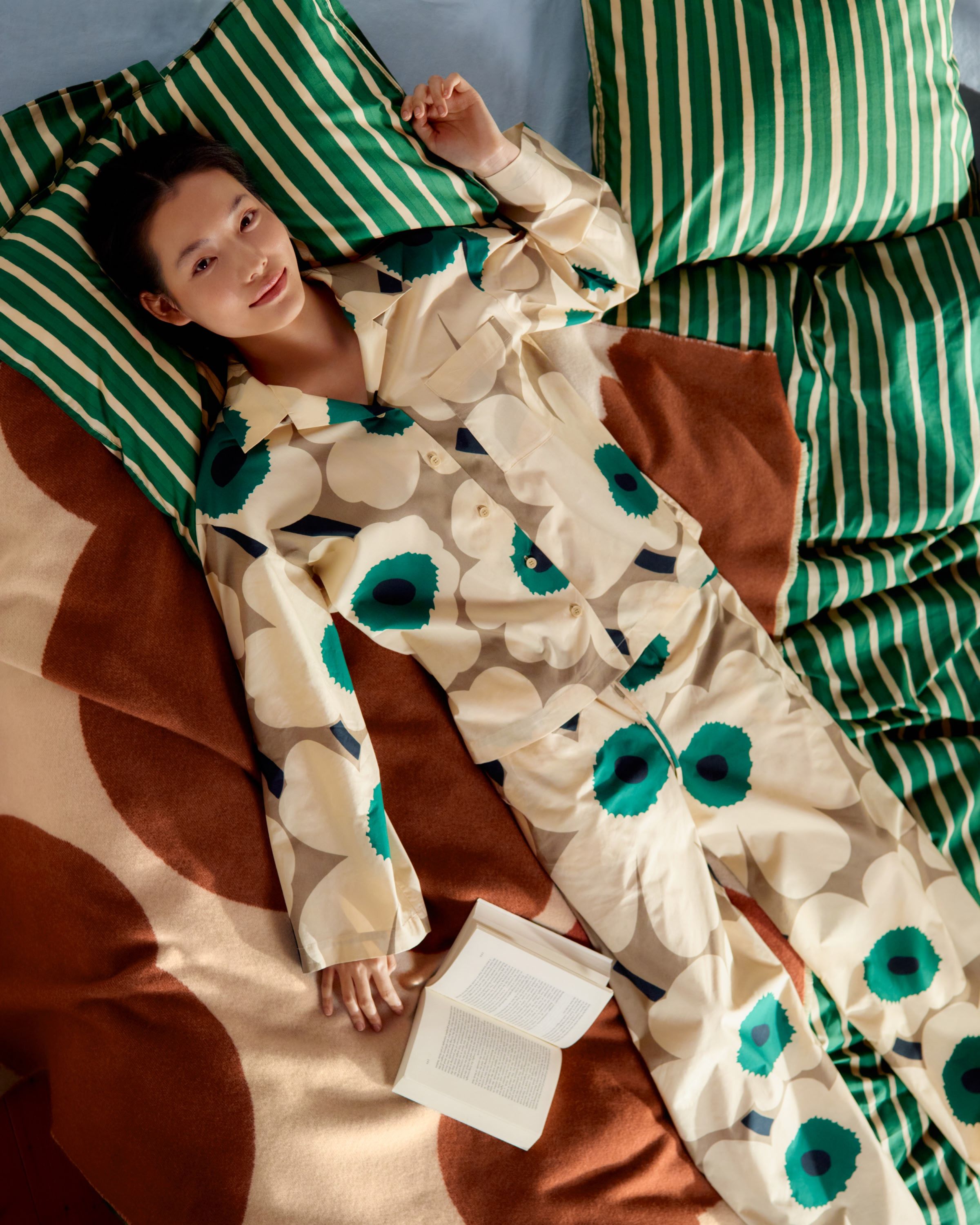 Model wearing an Unikko-print pyjama set, lying on a bed with Iso Piccolo-print bedlinen.