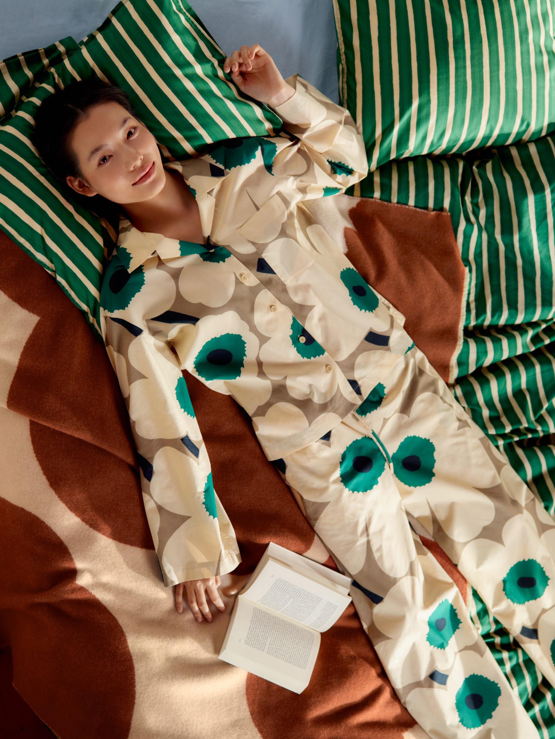 Model wearing an Unikko-print pyjama set, lying on a bed with Iso Piccolo-print bedlinen.