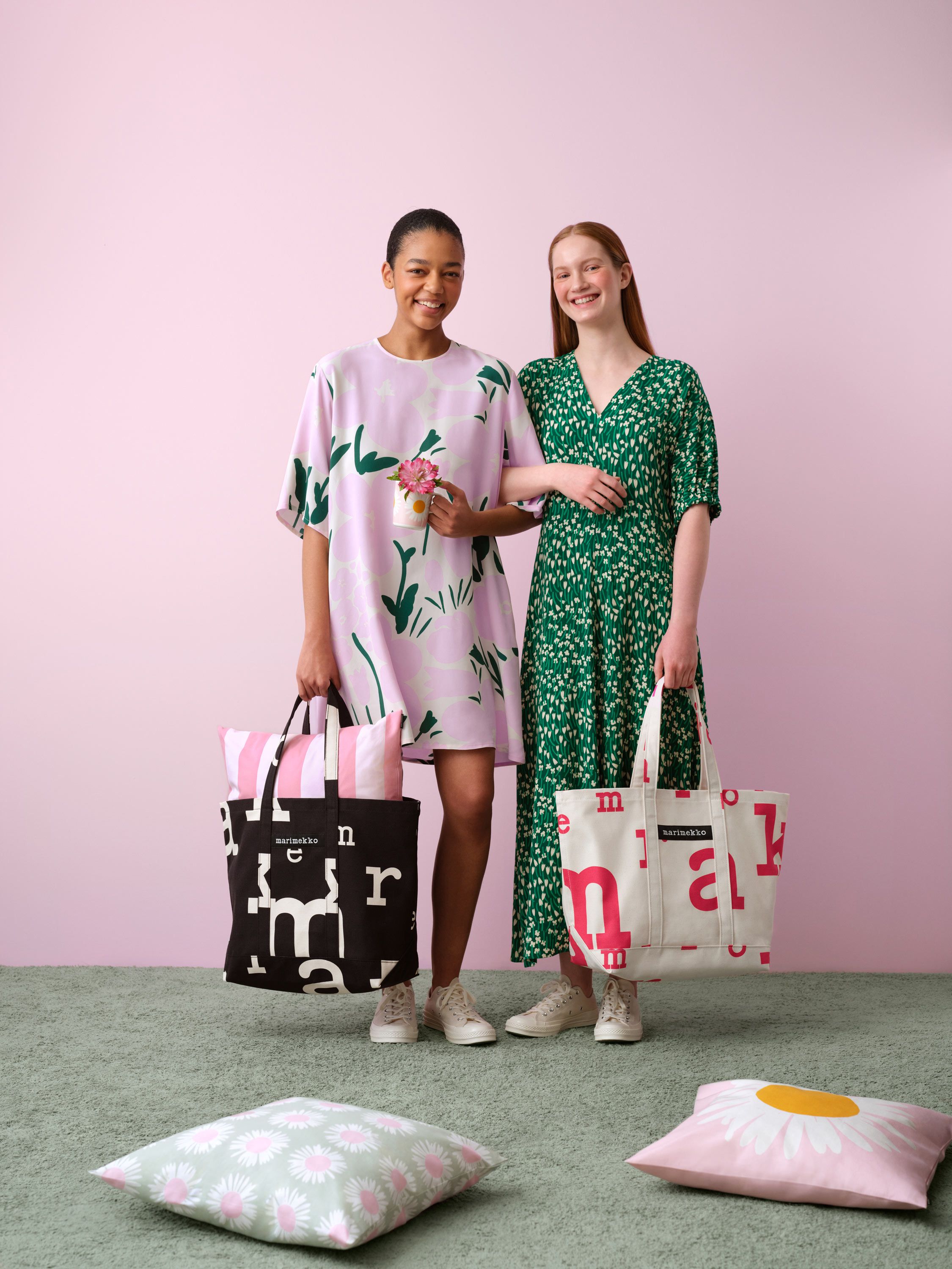 Mallien yllä mekot Tuulahdus- ja Talkoot-kuoseissa ja he pitelevät laukkuja, joissa on Marimerkki-kuosi. Maassa kaksi Auringonkukka-kuosi tyynyä.