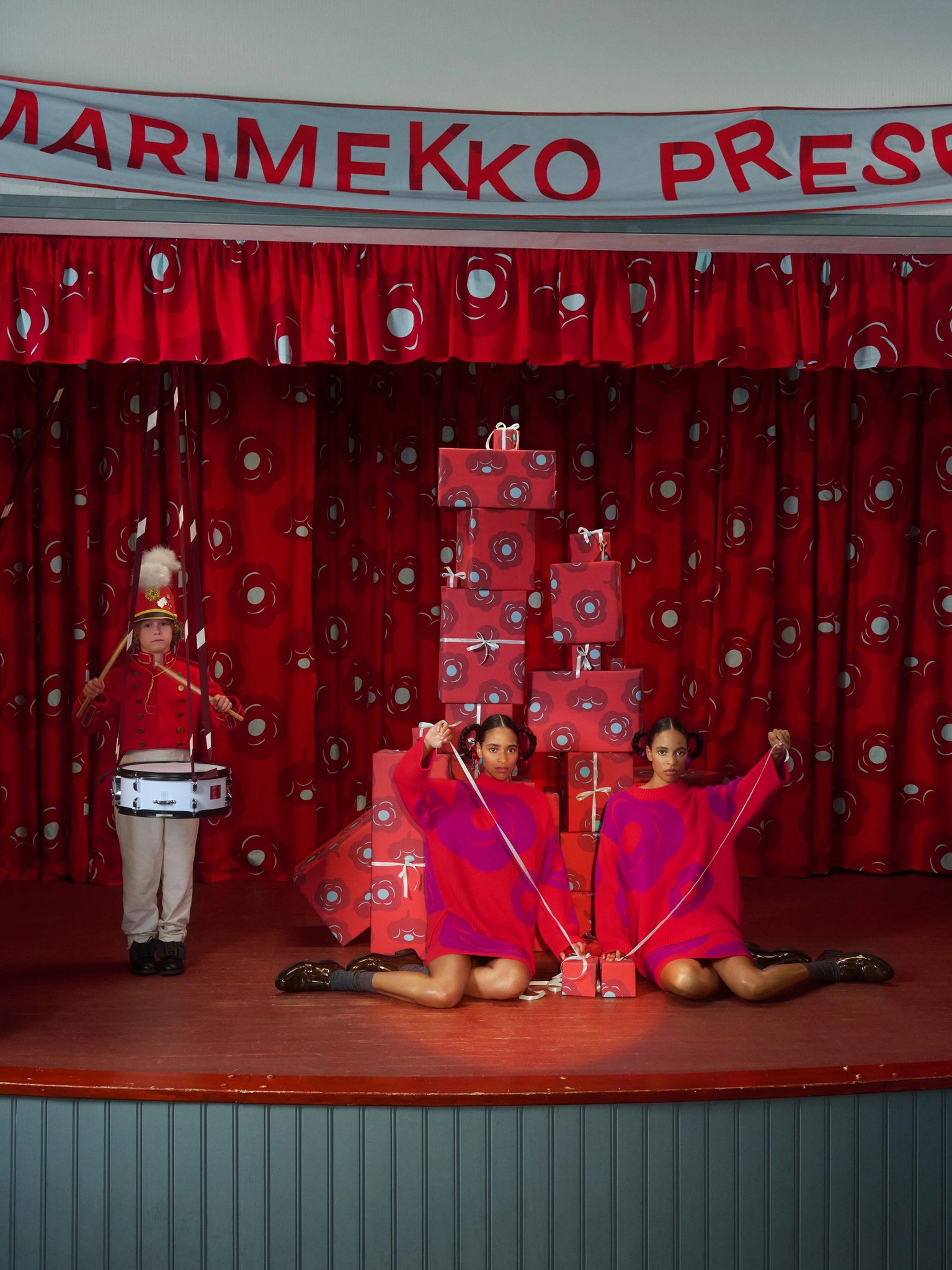Kaksi mallia istumassa ja poika soittamassa rumpua lavalla.