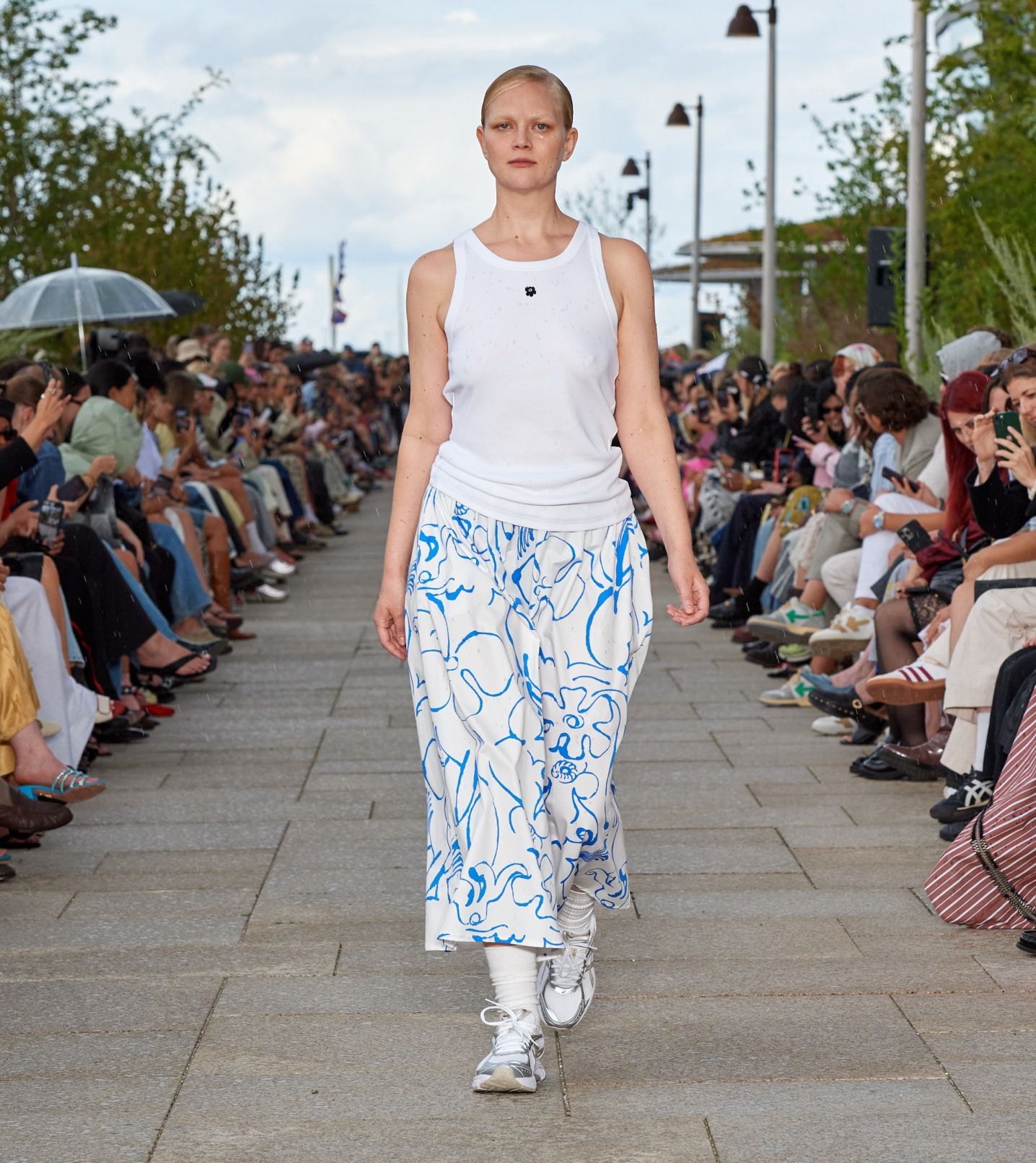 A model walking on the catwalk of a fashion show wearing a sleeveless white top and a white-blue skirt.
