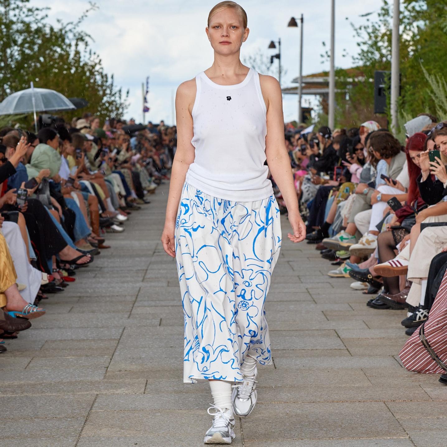 A model walking on the catwalk of a fashion show wearing a sleeveless white top and a white-blue skirt.