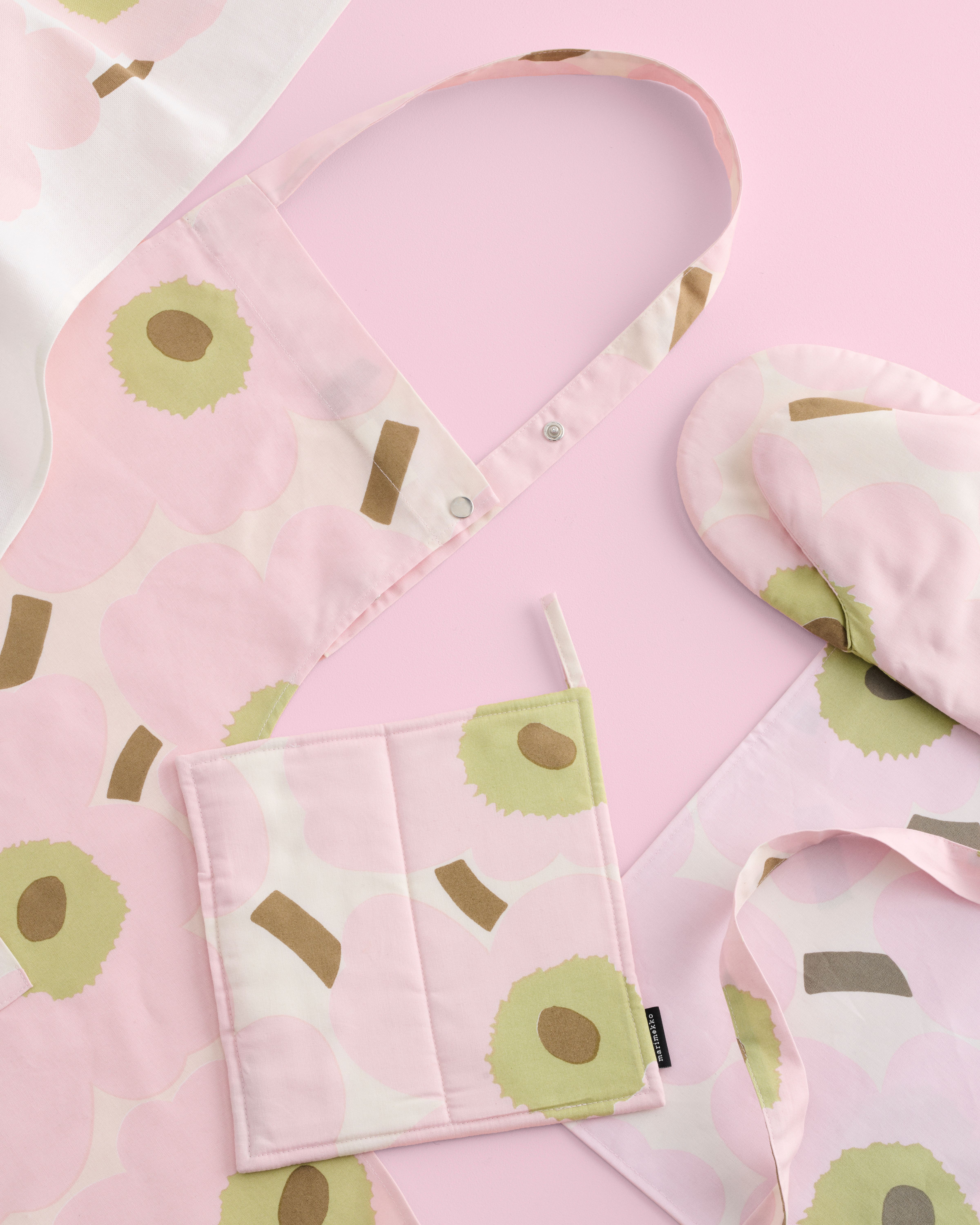Pink Unikko apron, oven mitts and pot holder displayed on a pink background
