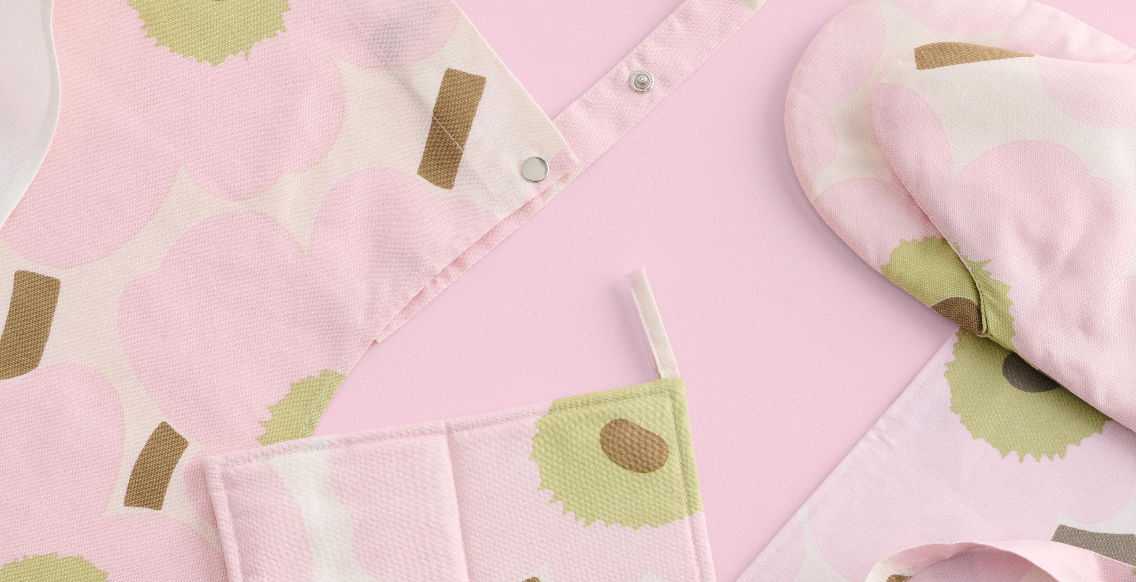 Pink Unikko apron, oven mitts and pot holder displayed on a pink background