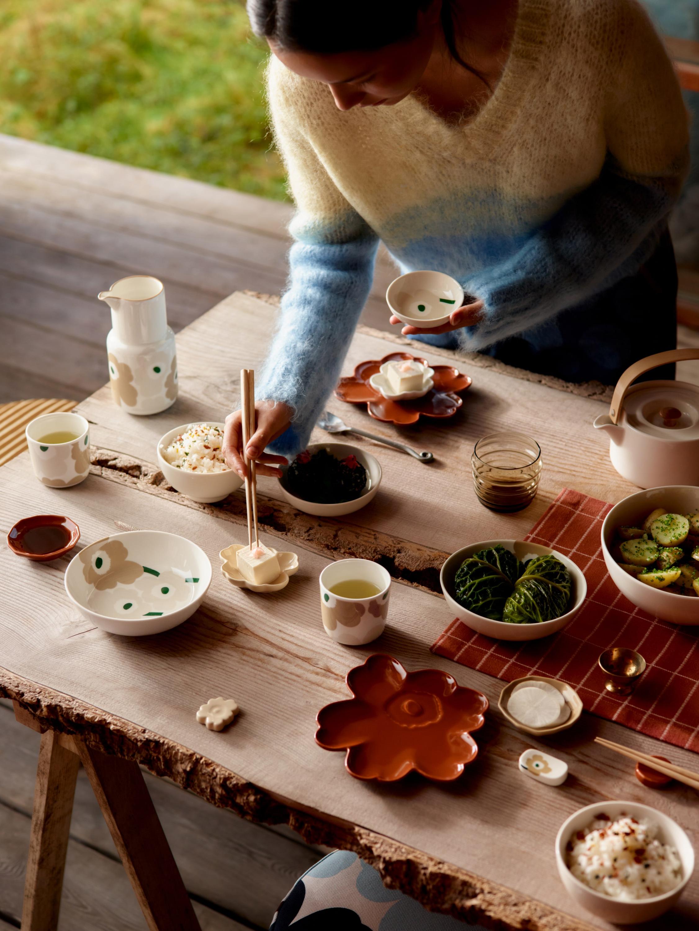 Model picking food with chopsticks from a table set with beige and red Unikko-print tableware.