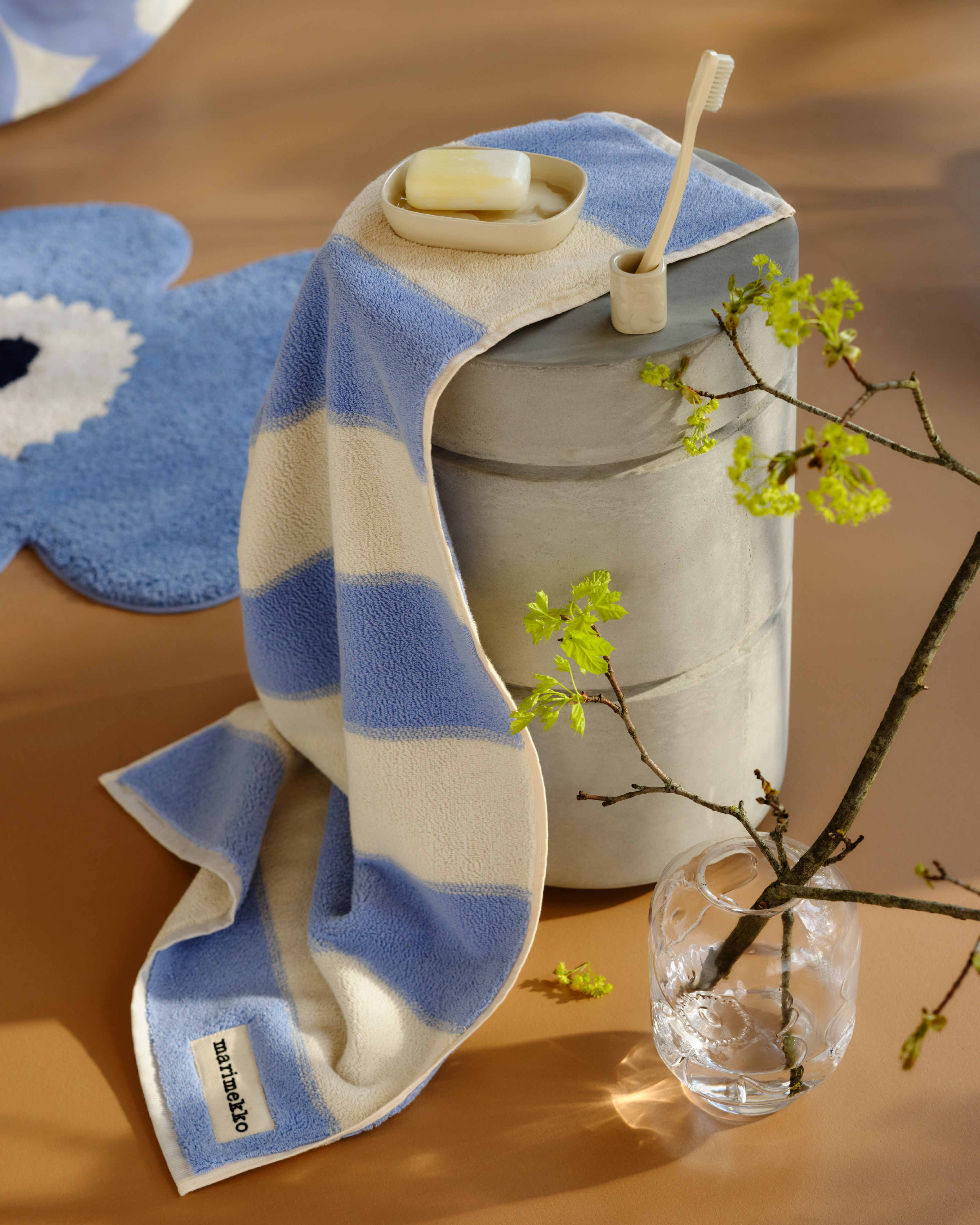 Blue and white striped towel, blue Unikko-shaped bath mat and a glass vase with a twig on the floor