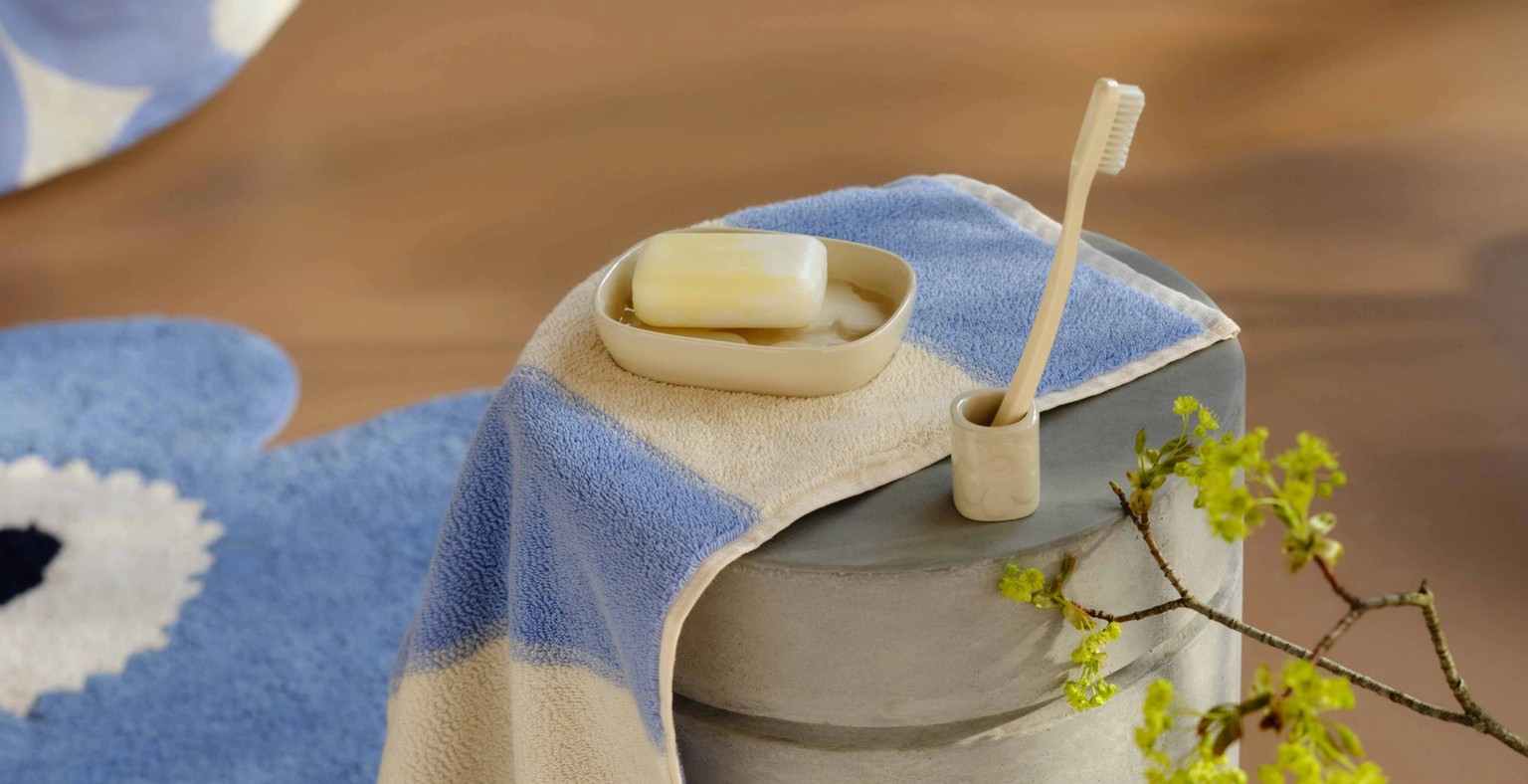 Blue and white striped towel, blue Unikko-shaped bath mat and a glass vase with a twig on the floor