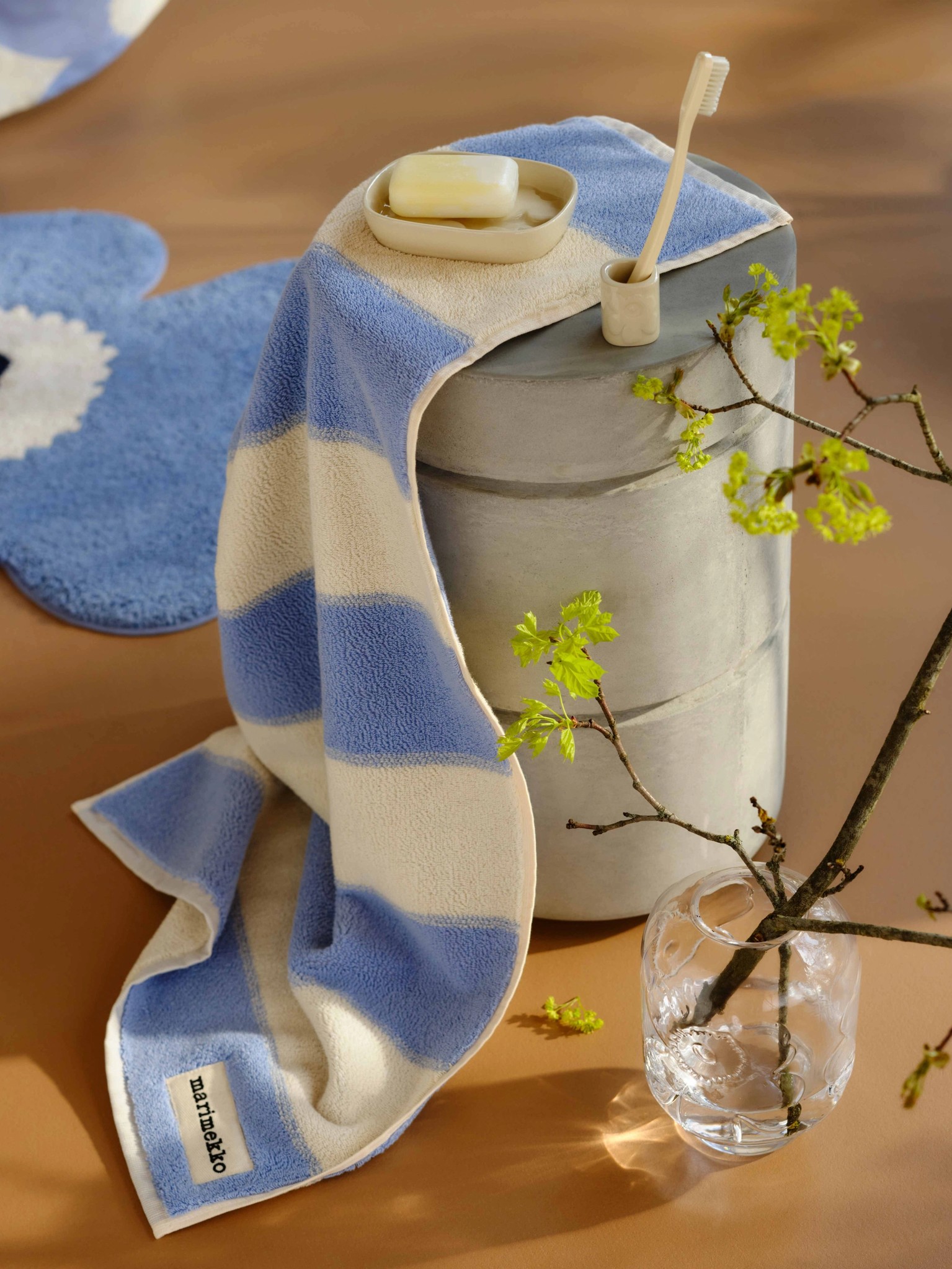 Blue and white striped towel, blue Unikko-shaped bath mat and a glass vase with a twig on the floor