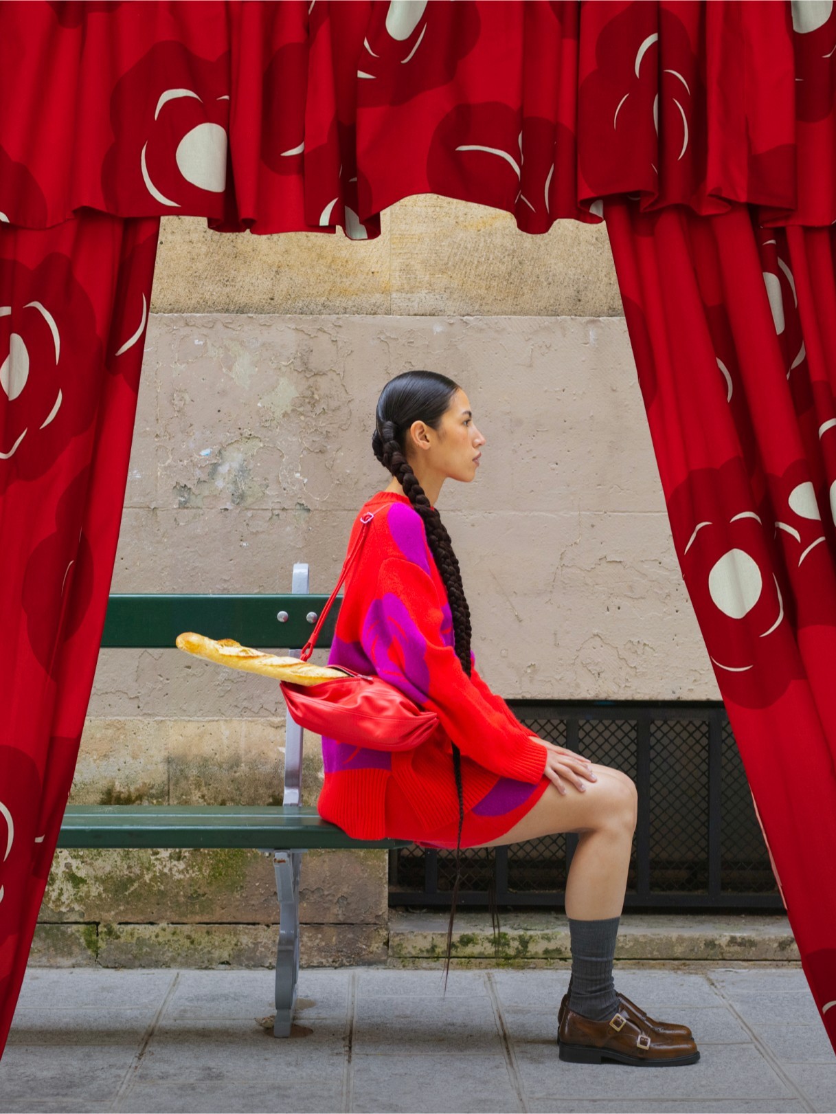 A view between red printed curtains of a model sitting on a bench with a baguette in her bag.