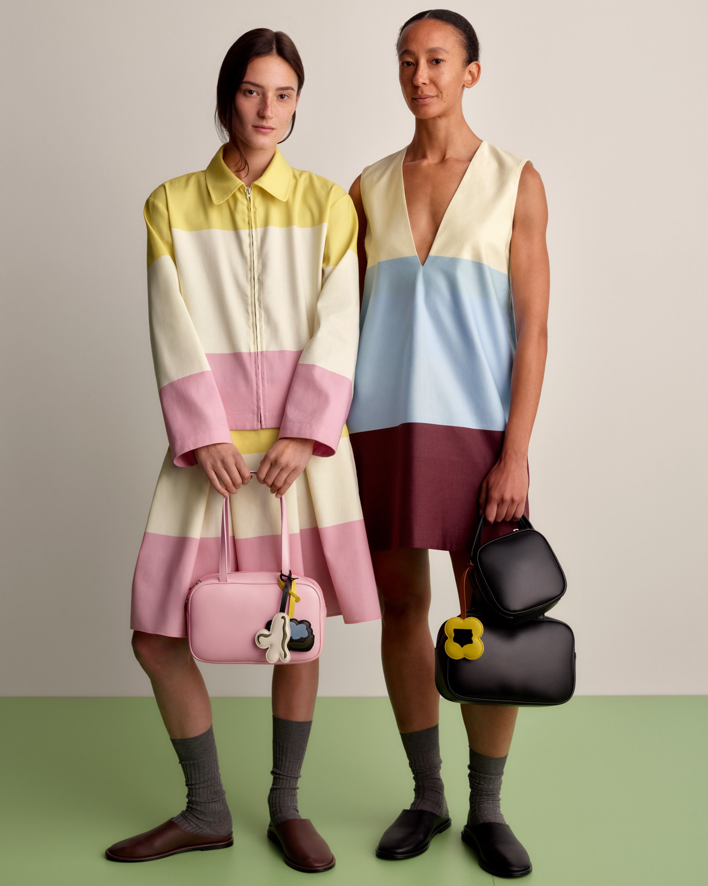 One model wearing a collar shirt and matching skirt in Hennika print, and another model wearing a sleeveless Hennika dress in darker colors. Both models carry Gratha leather bags in two different sizes and colors.