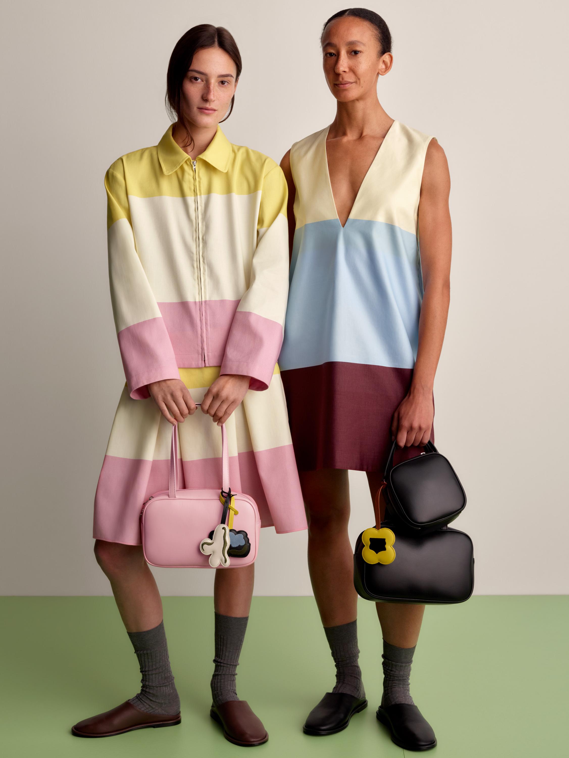 One model wearing a collar shirt and matching skirt in Hennika print, and another model wearing a sleeveless Hennika dress in darker colors. Both models carry Gratha leather bags in two different sizes and colors.