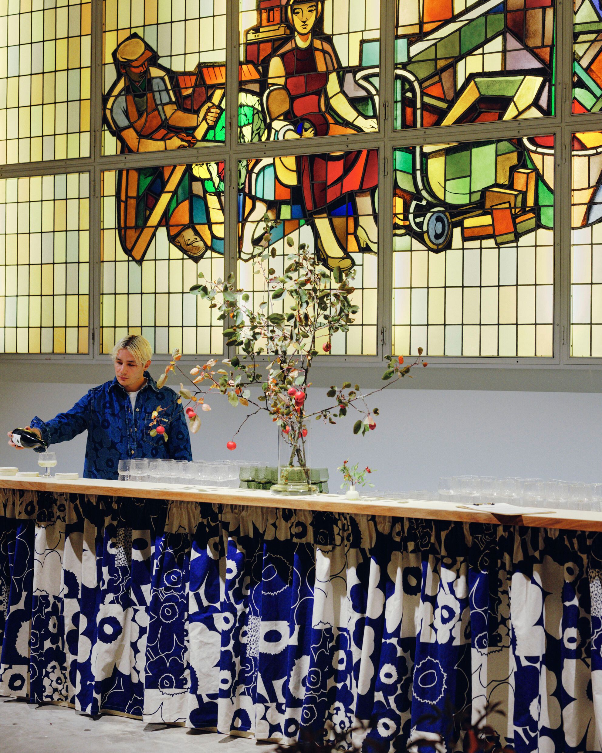 A waiter pouring wine at a bar counter, which is decorated with a blue and white Unikko-patterned curtain. In the background, a window with mosaic glass art.