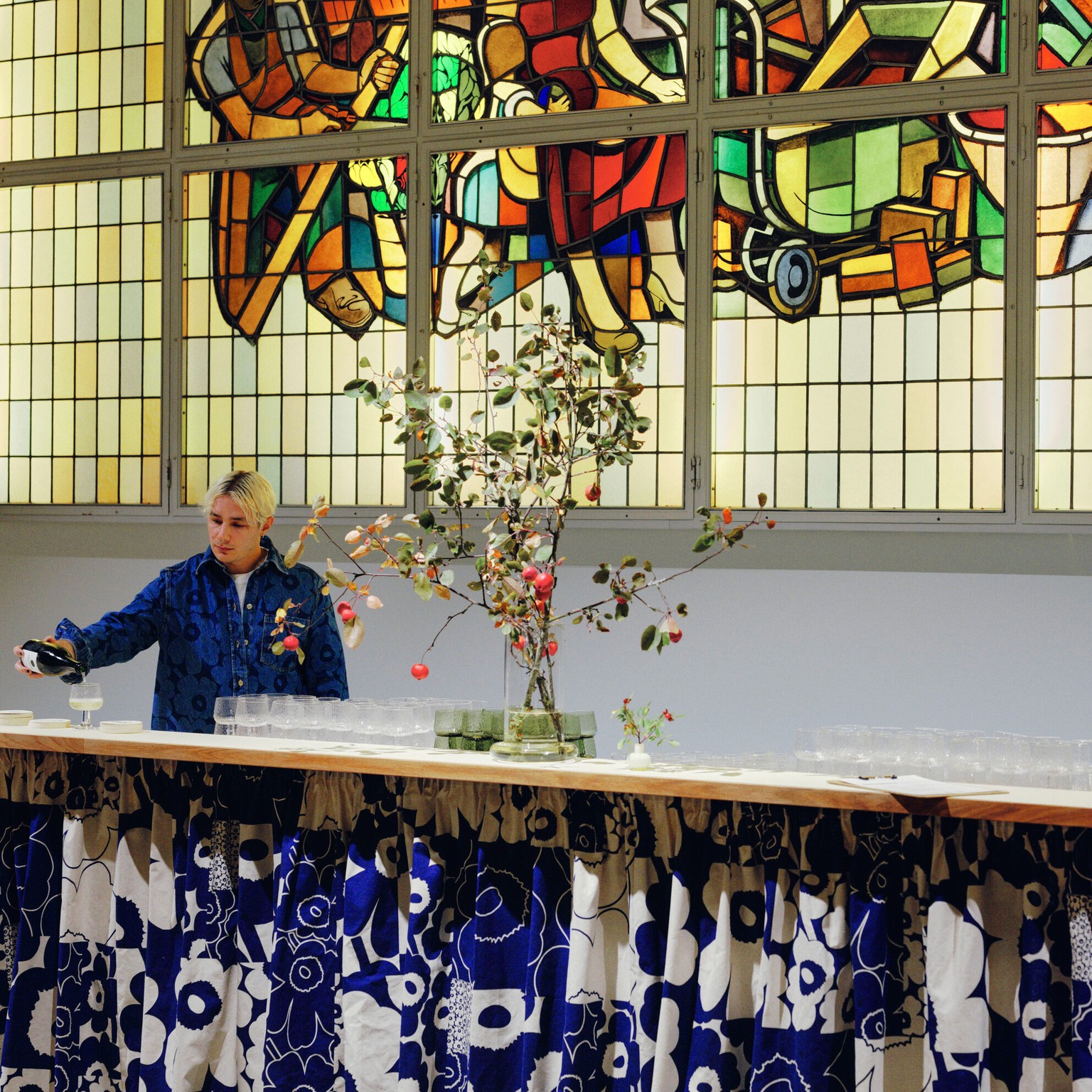 A waiter pouring wine at a bar counter, which is decorated with a blue and white Unikko-patterned curtain. In the background, a window with mosaic glass art.