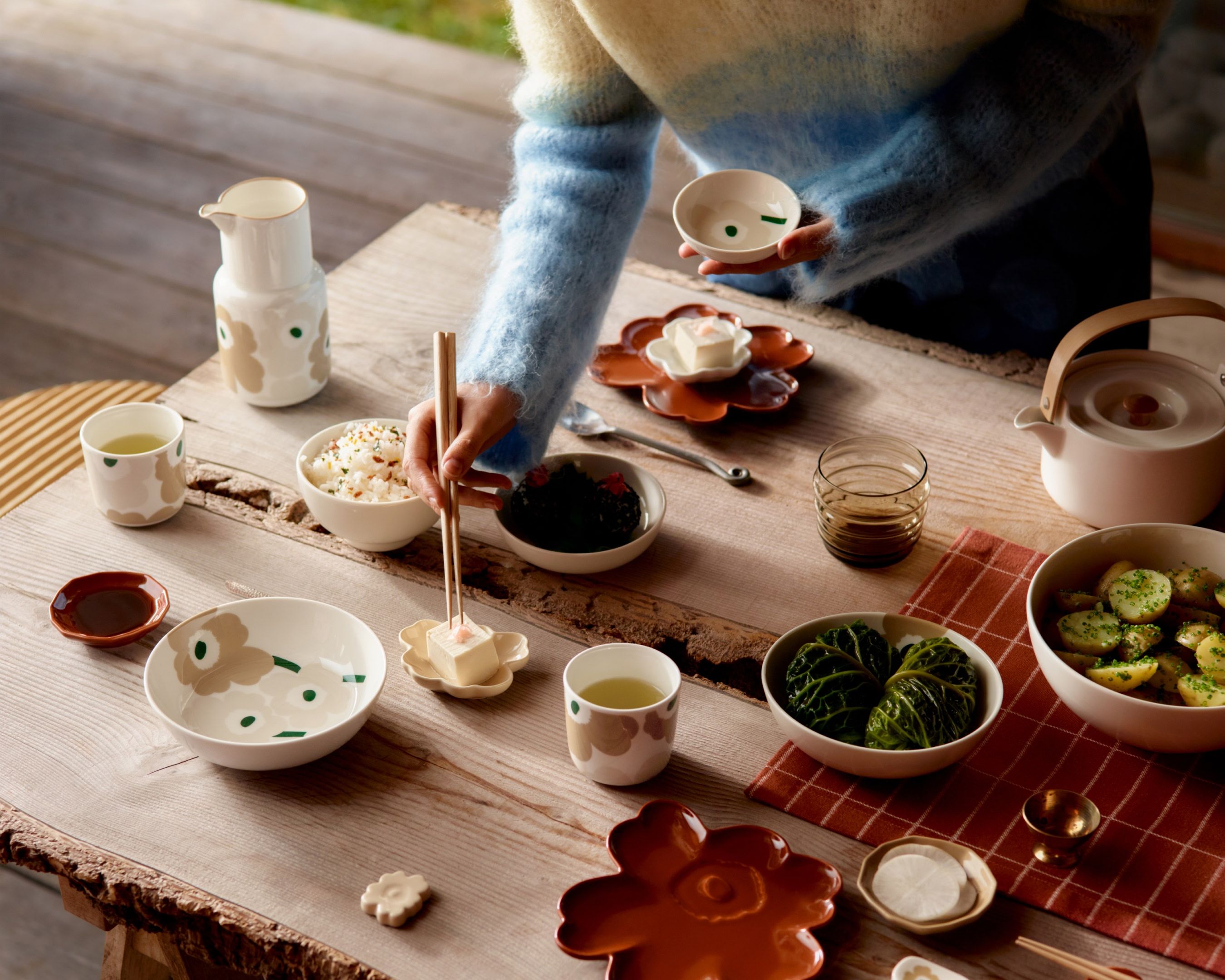 Model picking food with chopsticks from a table set with beige and red Unikko-print tableware.