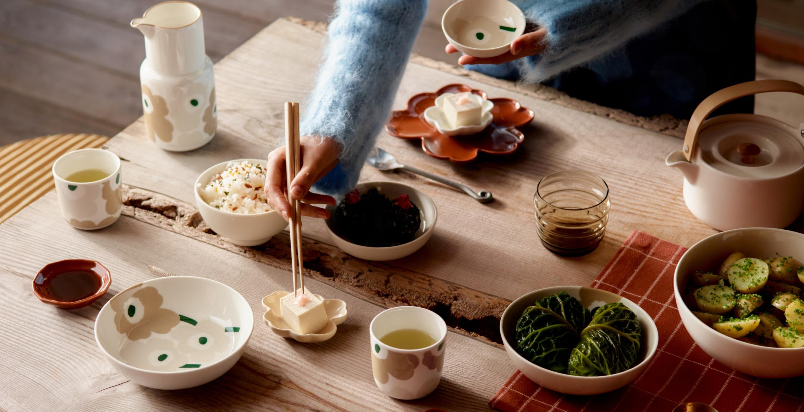 Model picking food with chopsticks from a table set with beige and red Unikko-print tableware.