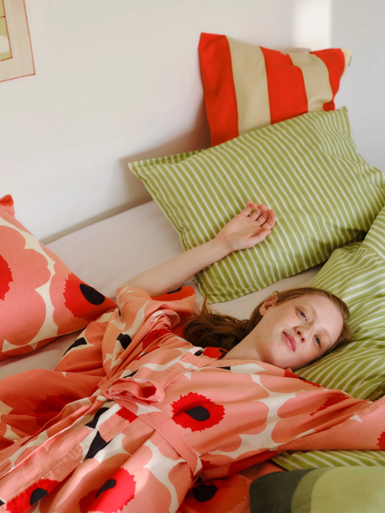 Two pictures, one with red and white Unikko towels next to the washbasin and the other with the model lying on the bed in a Unikko bathrobe with green Piccolo sheets.