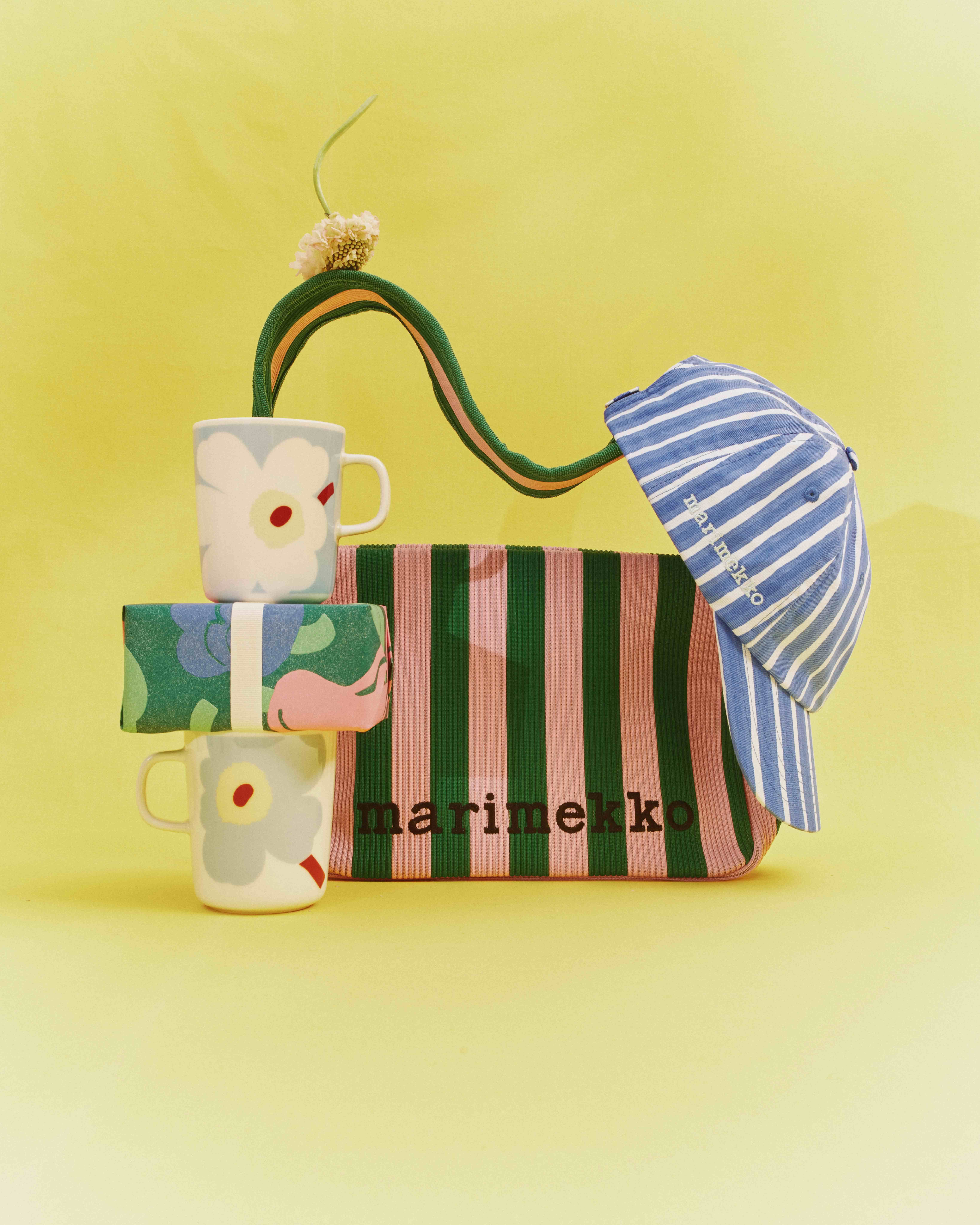 Marimekko's Unikko mugs stacked on top of a gift box; behind the mugs is a Knitted Shoulderbag bag with a blue-and-white beanie hanging from it, all arranged against a yellow background.