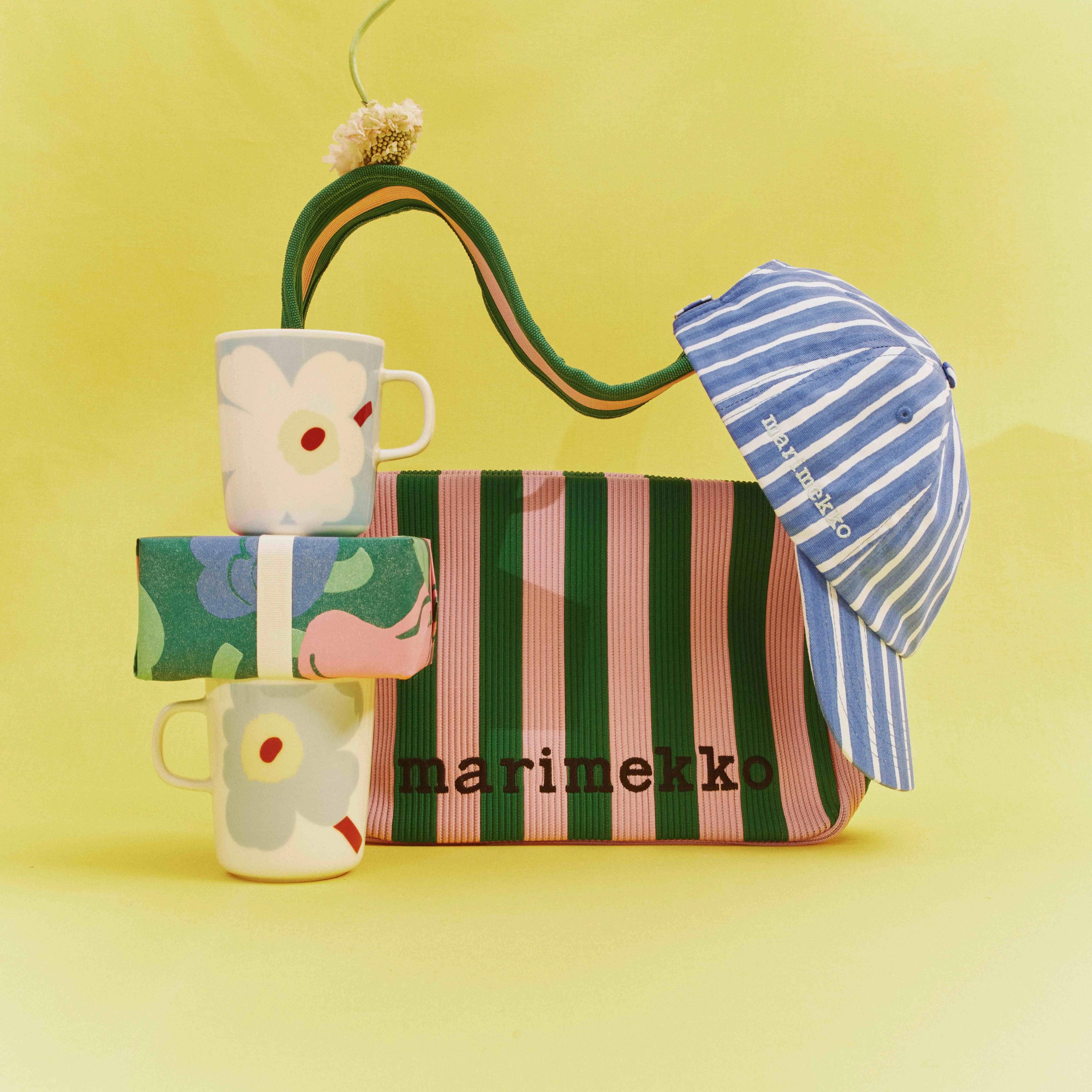 Marimekko's Unikko mugs stacked on top of a gift box; behind the mugs is a Knitted Shoulderbag bag with a blue-and-white beanie hanging from it, all arranged against a yellow background.