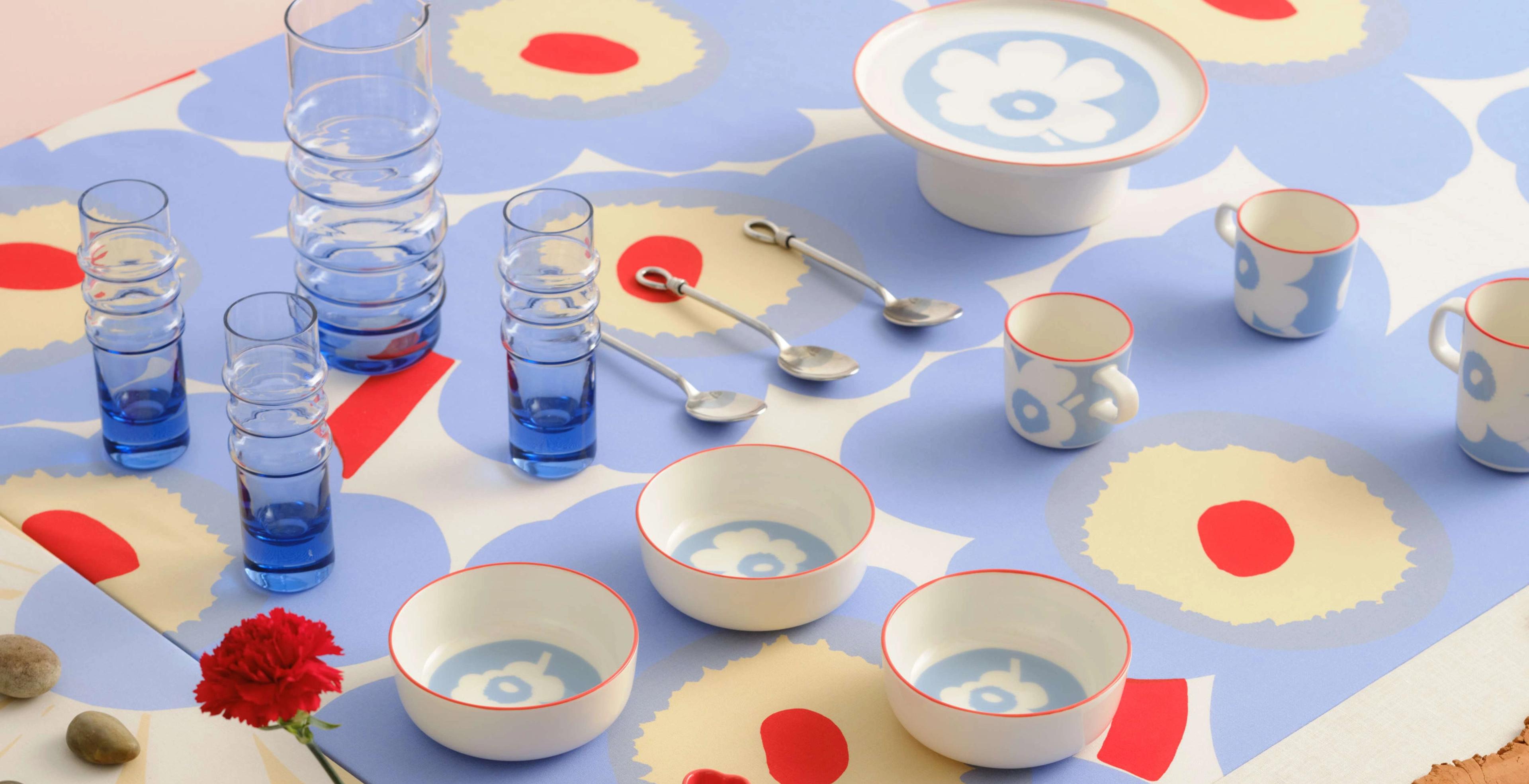 Various Marimekko glassware and tableware in Unikko and Piccolo prints, including plates, bowls, mugs, espresso cups, and coasters, arranged in rows on a large table