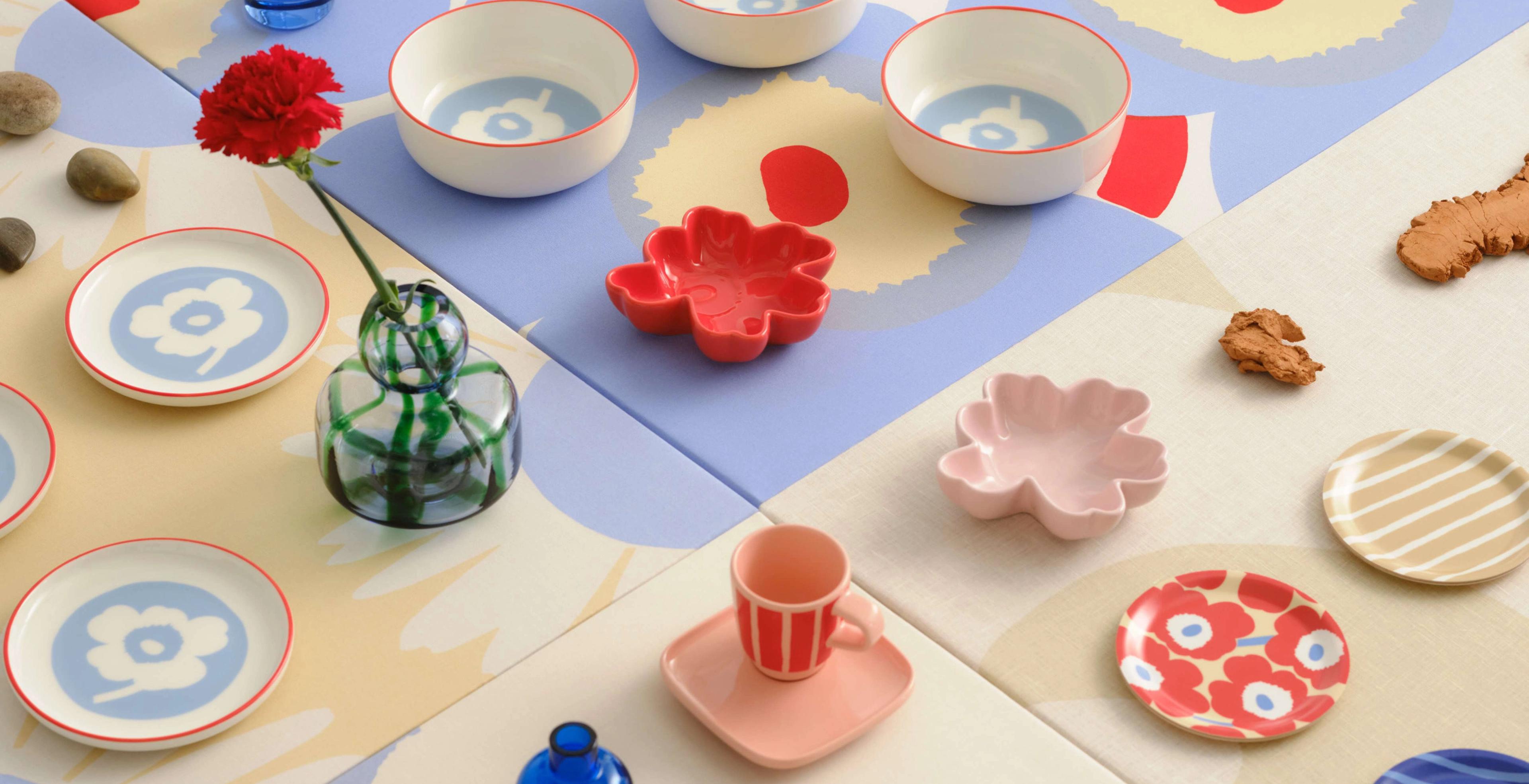 Various Marimekko glassware and tableware in Unikko and Piccolo prints, including plates, bowls, mugs, espresso cups, and coasters, arranged in rows on a large table