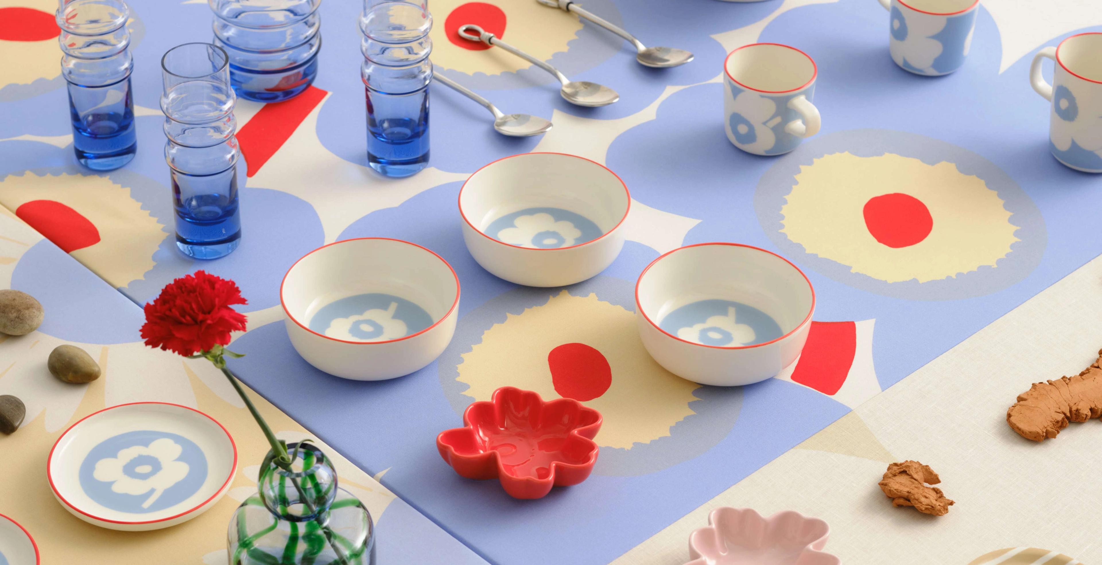 Various Marimekko glassware and tableware in Unikko and Piccolo prints, including plates, bowls, mugs, espresso cups, and coasters, arranged in rows on a large table