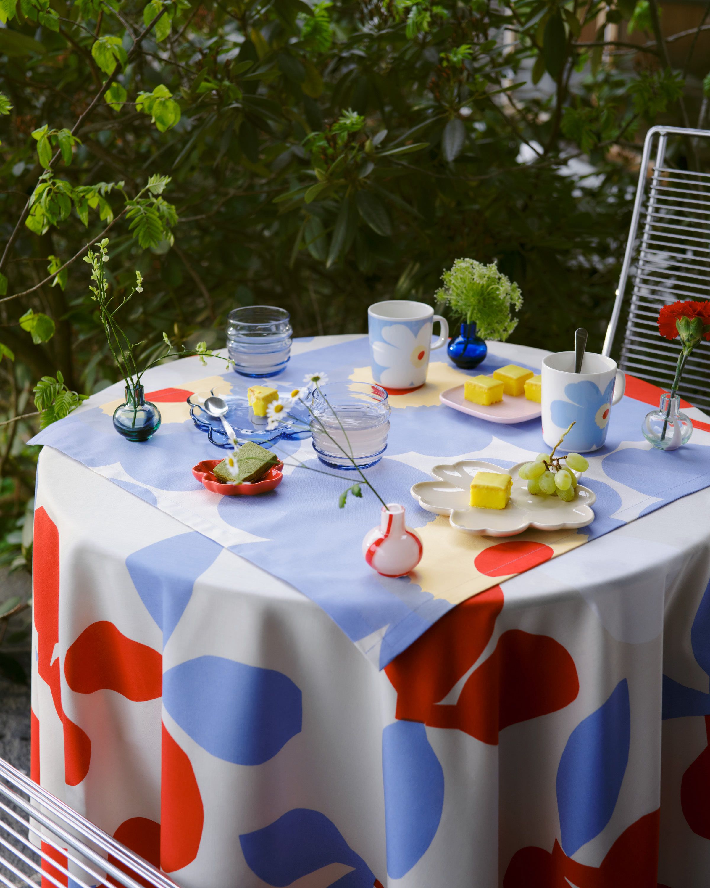 Outdoor table setting with blue Sukat Makkaralla glasses, Unikko-shaped plates, mini vases, and a Pieni Omppu print tablecloth.