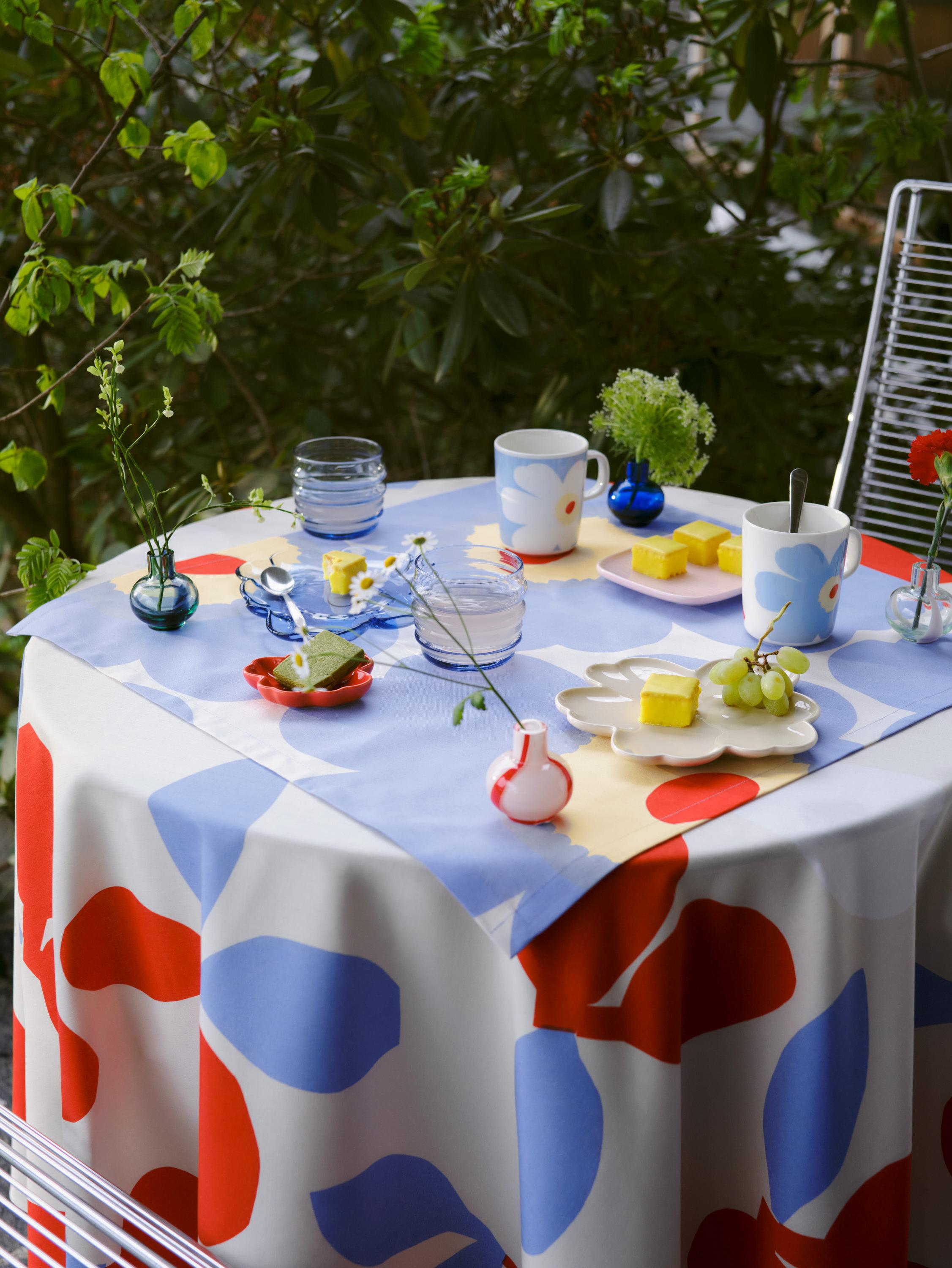 Outdoor table setting with blue Sukat Makkaralla glasses, Unikko-shaped plates, mini vases, and a Pieni Omppu print tablecloth.