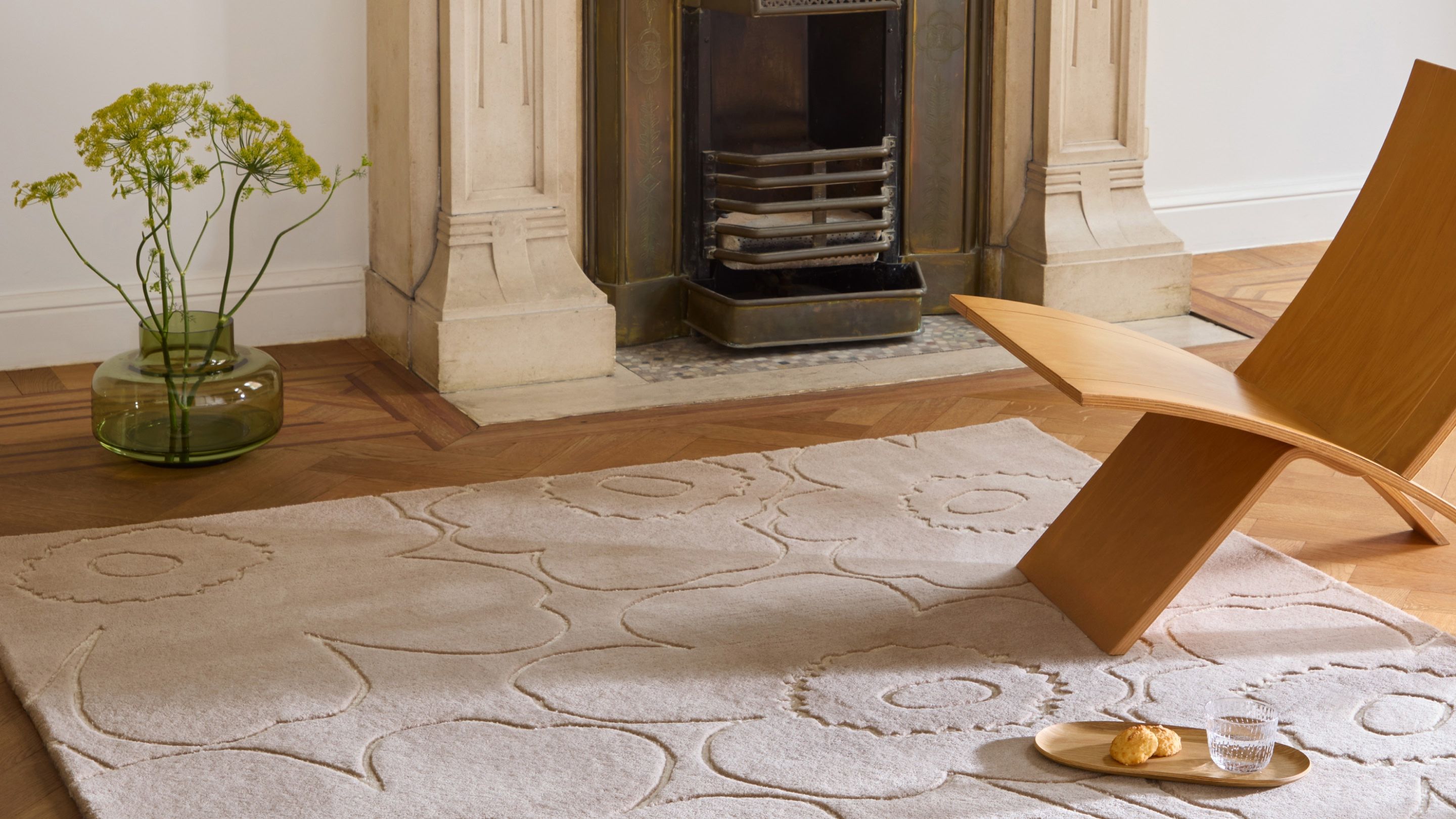 Beige rug with Piirto Unikko pattern in front of a fireplace and Urna vase on the side with flowers.